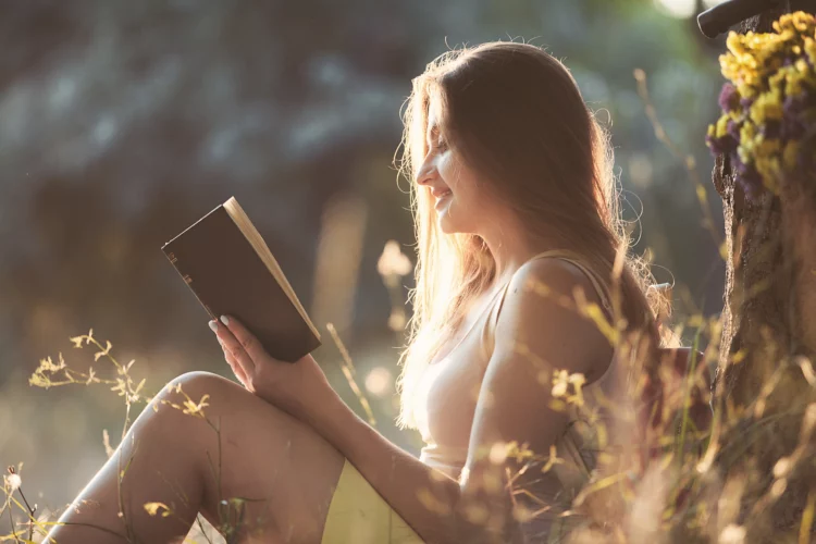 Beautiful young lady sitting in a park under a tree reading a book