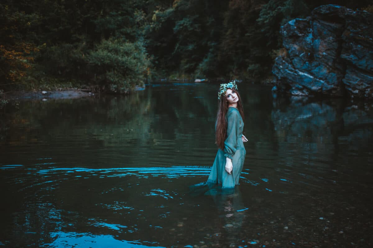 Young woman walking through the river in a dress with flower wreath on her head