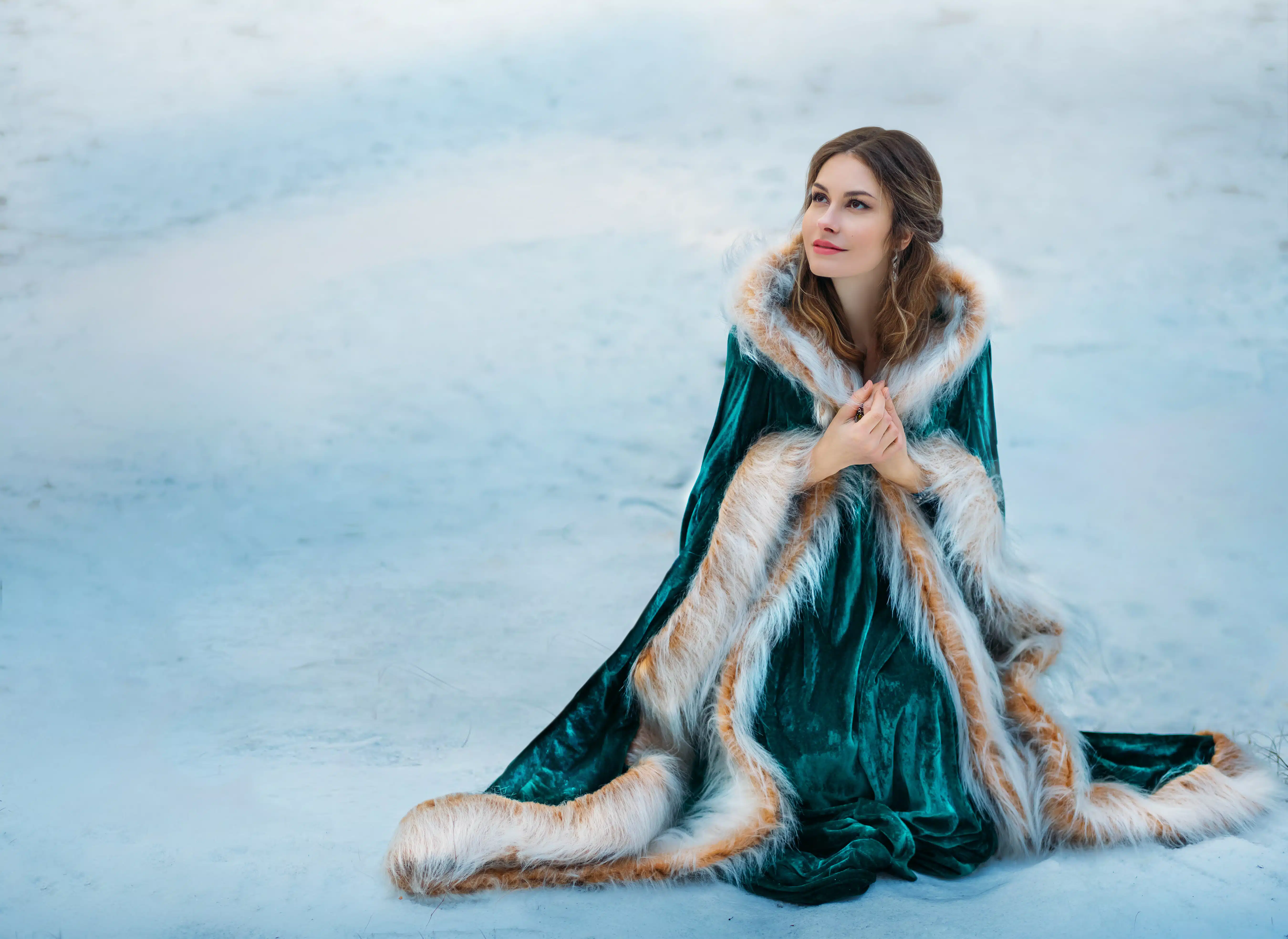 Fantasy woman sits on white snow in winter forest. Princess girl smiling face. Green long velvet vintage coat fur. Mystical image of frozen lady wanderer.
