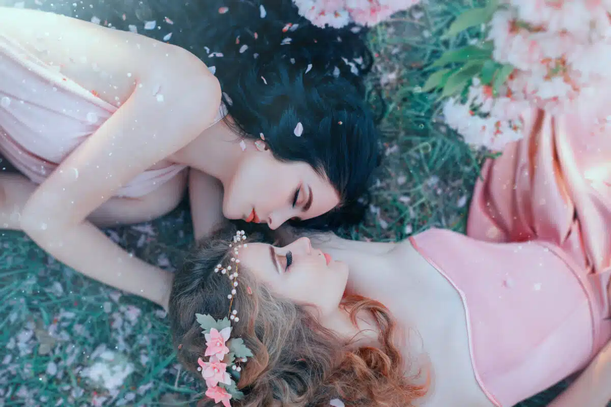 portrait two young women lie cuddling on grass in spring garden, luxurious long curly hair strewn with flowers rose petals. Attractive face gentle makeup. Different sisters. Natural cosmetics concept