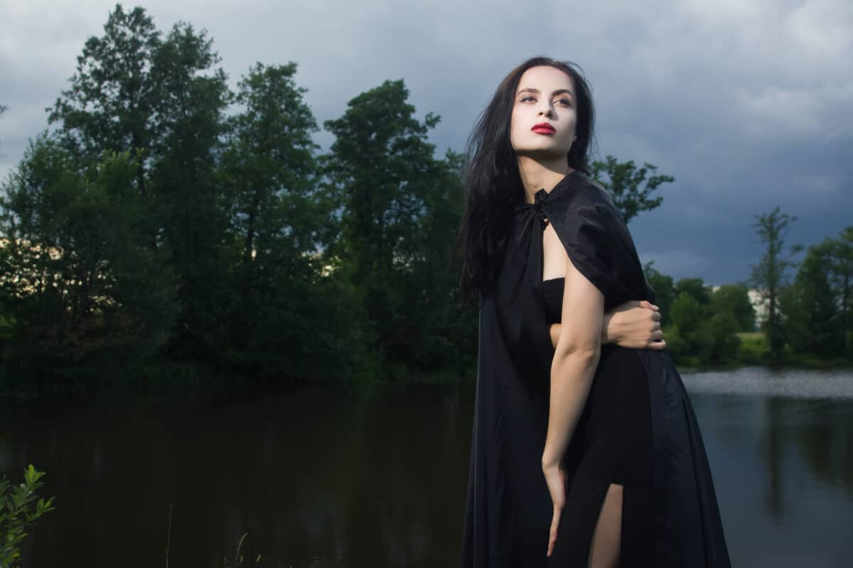 a brunette woman in a black dress and black cloak standing by the lake