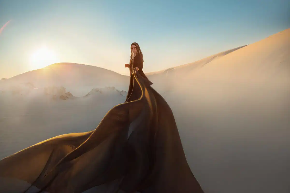 Mystery arabic woman in black long dress stands in desert long train silk fabric fly flytter in wind motion. clothes gold accessories hide face. Oriental fashion model. Sand dunes background sunset.