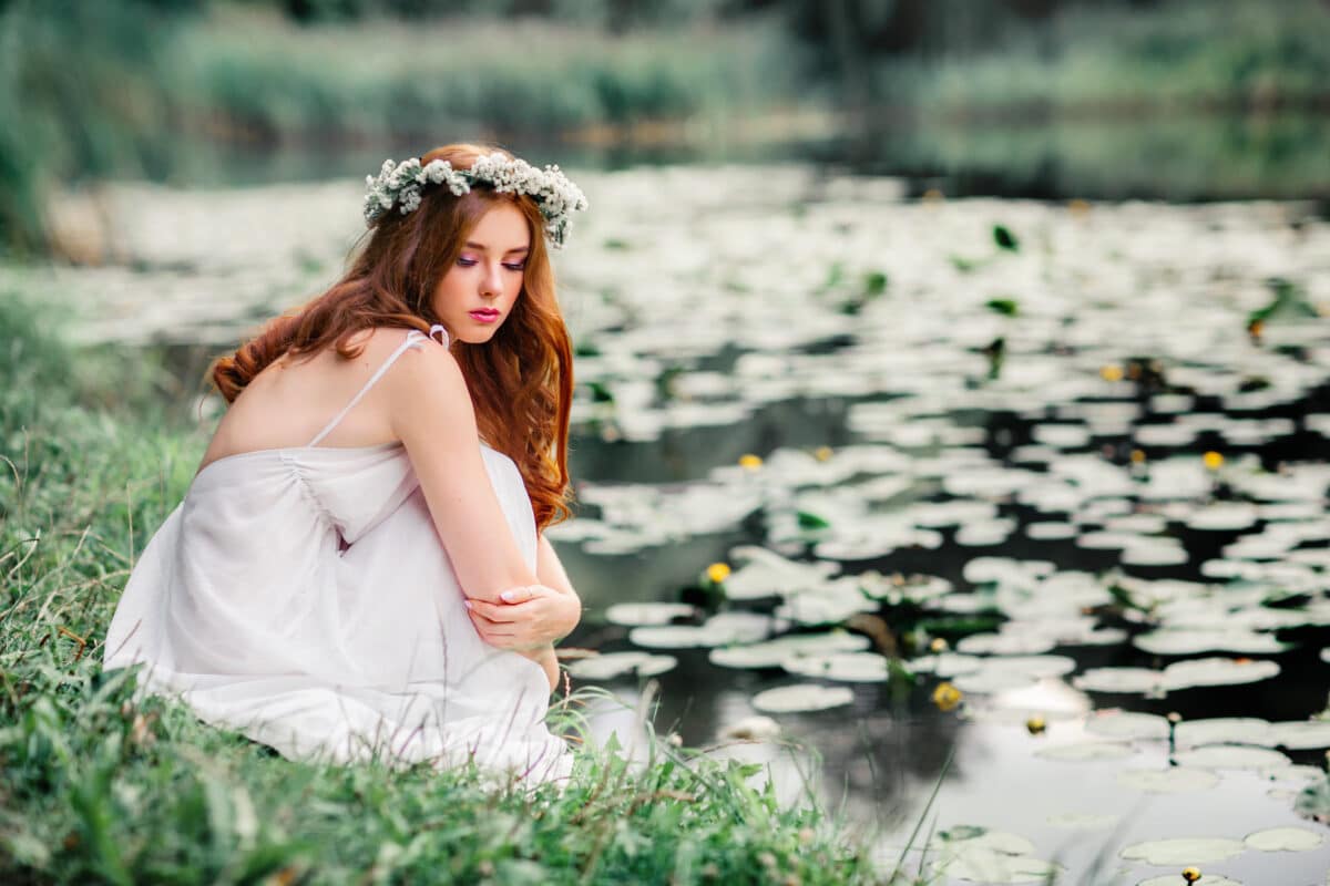 Beautiful red haired girl in white dress and wreath of wild flowers sitting by the pond with wild water lilies