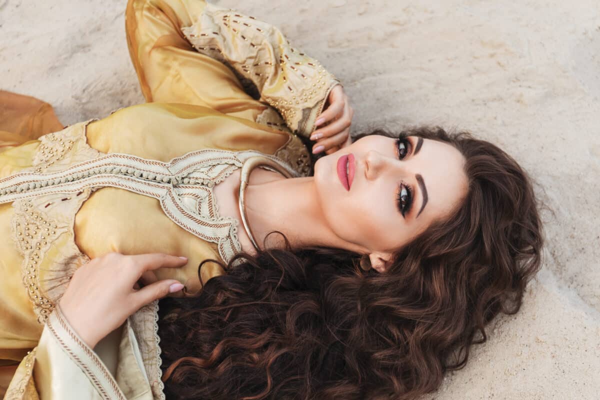 beautiful sexy young brunette woman with curly hair and with evening makeup, in golden evening dress in oriental style and with golden accessories lies in dessert sands on the beach