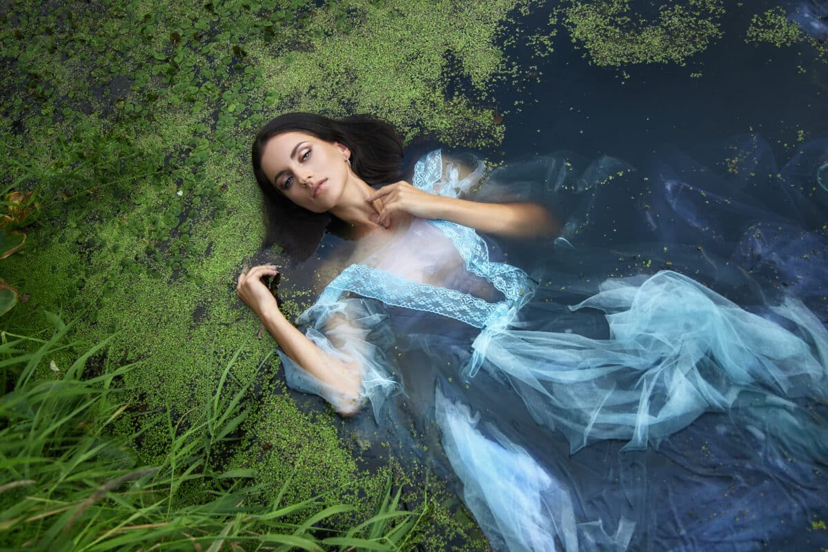 Art beautiful romantic woman lies in swamp in blue long dress with flowers. Portrait brunette in transparent dress in water swamp mud duckweed.