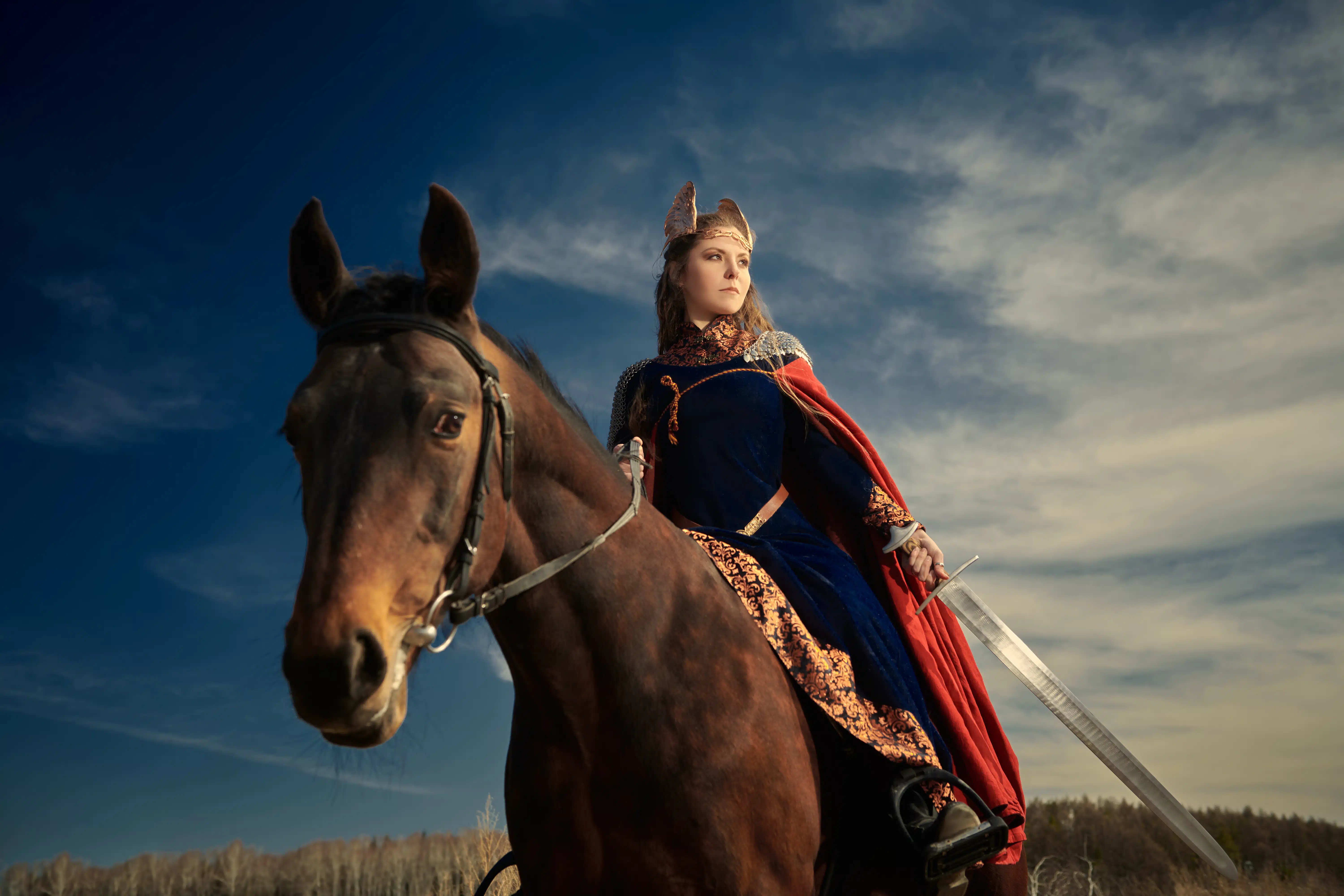 female medieval warrior holding a sword while riding a brown horse