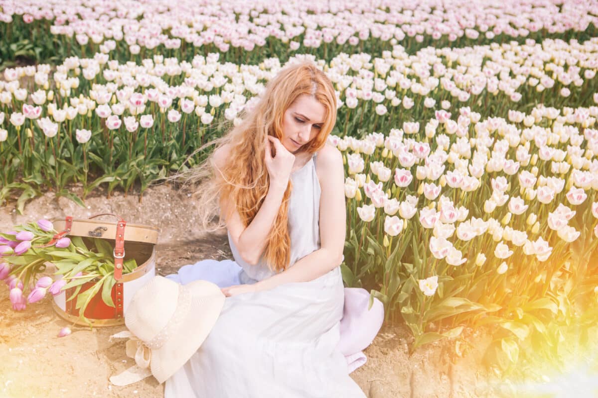 Beautiful redhead wearing a white dress sitting amid colorful tulip flower fields