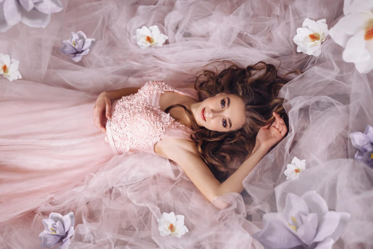 A young princess with long hair in a sumptuous pink dress lies on the floor with flowers scattered around her