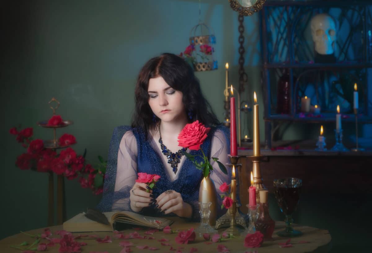 young beautiful but sad woman in blue vintage dress with book on table holding a red rose and staring at it