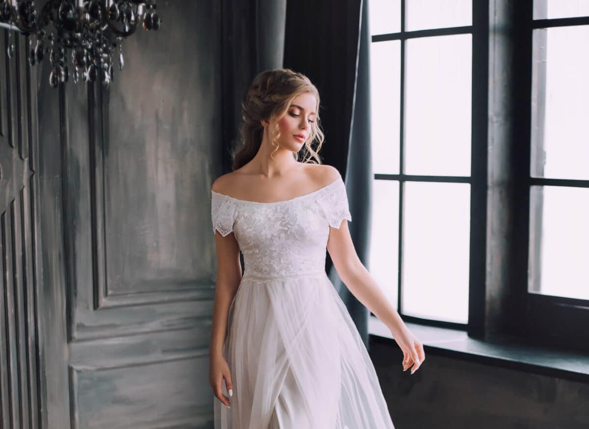mysterious pretty lady with blond curly hair looks down modestly, enchanted girl in chic light white long vintage dress with open shoulders in dark cloak, princess in gothic castle with large windows