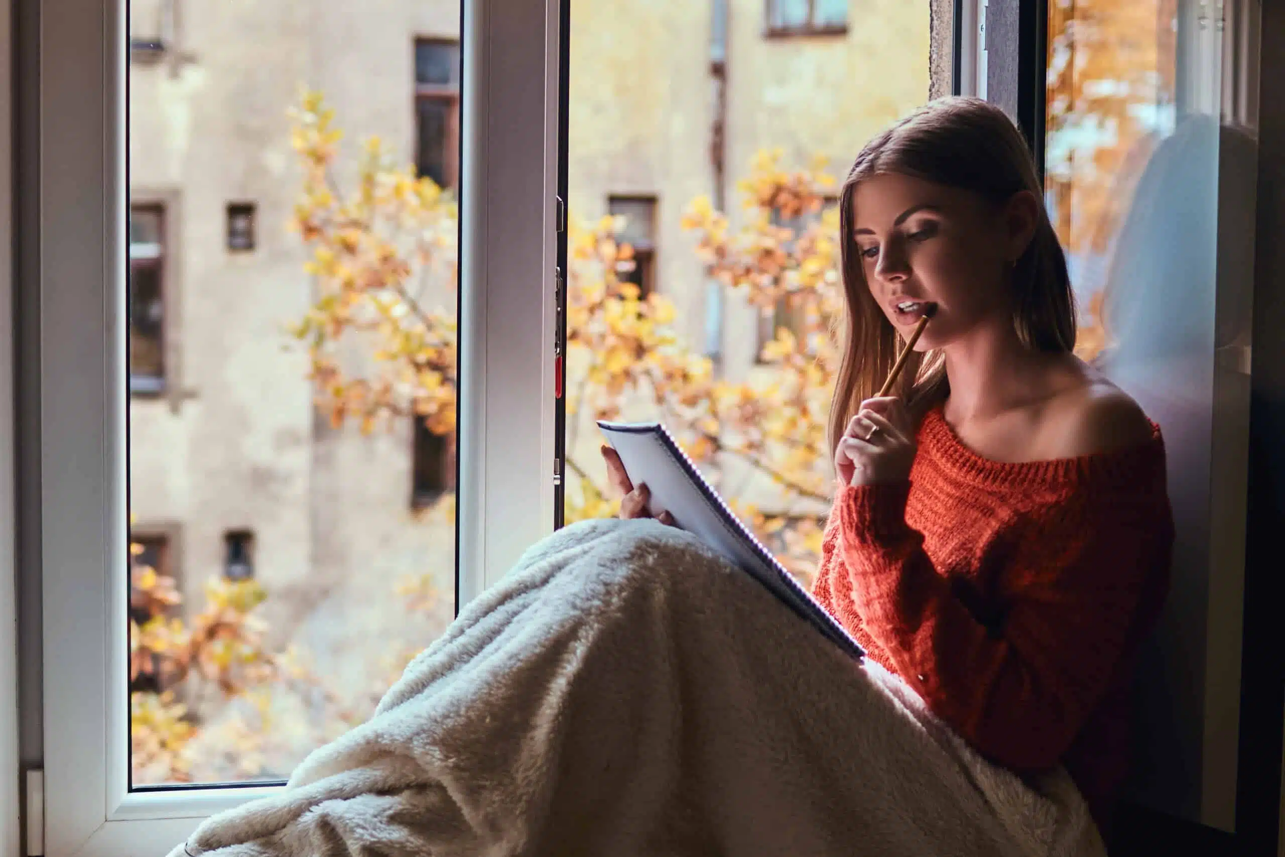 girl in a warm sweater covered her legs with a blanket, writing in her notebook sitting on the window sill