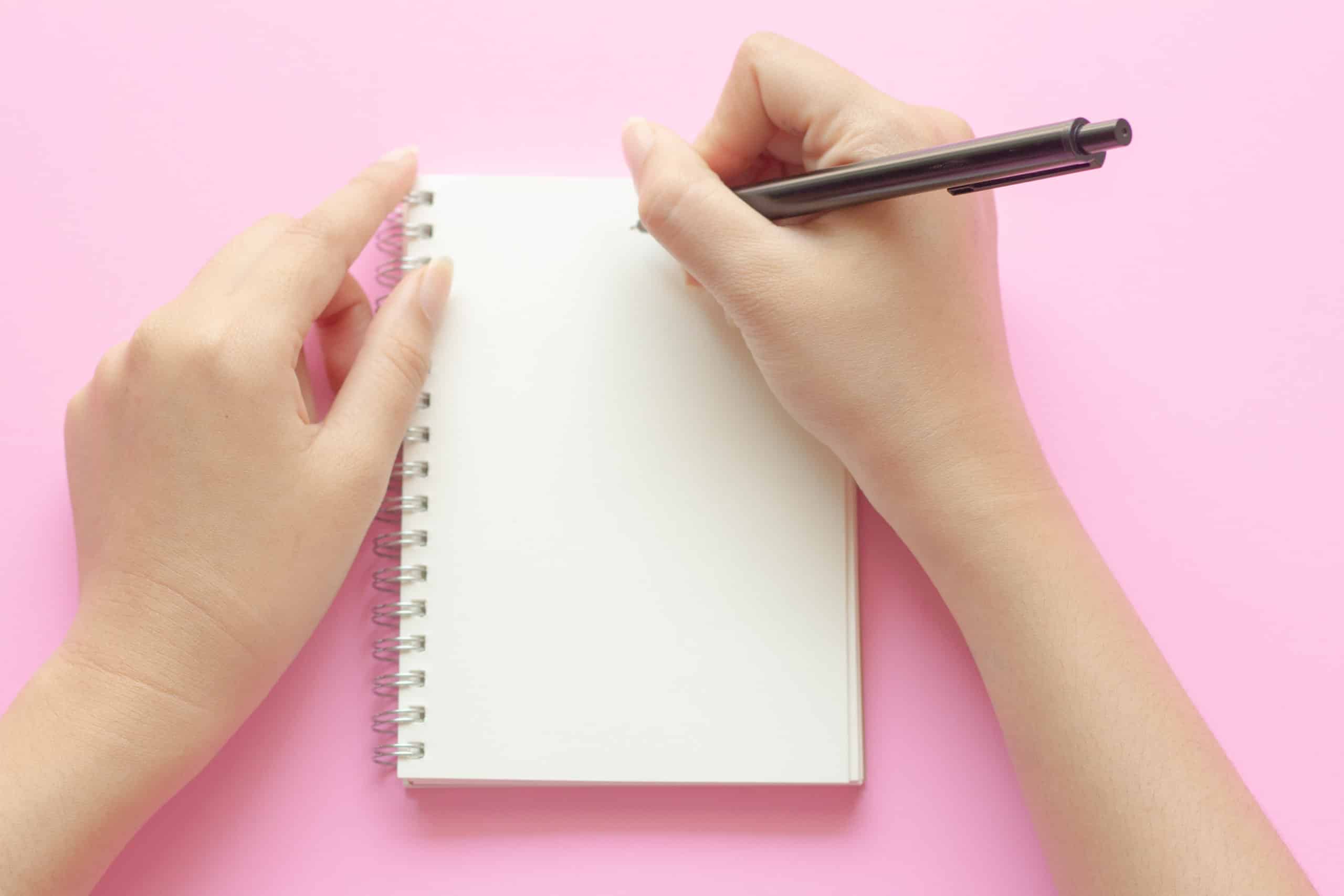 woman's hands writing in empty notebook at the pink desk.
