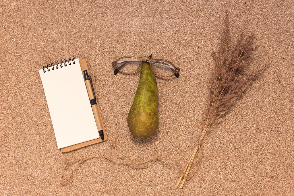 Blank notepad, glasses on pear and dry grass on cork table.