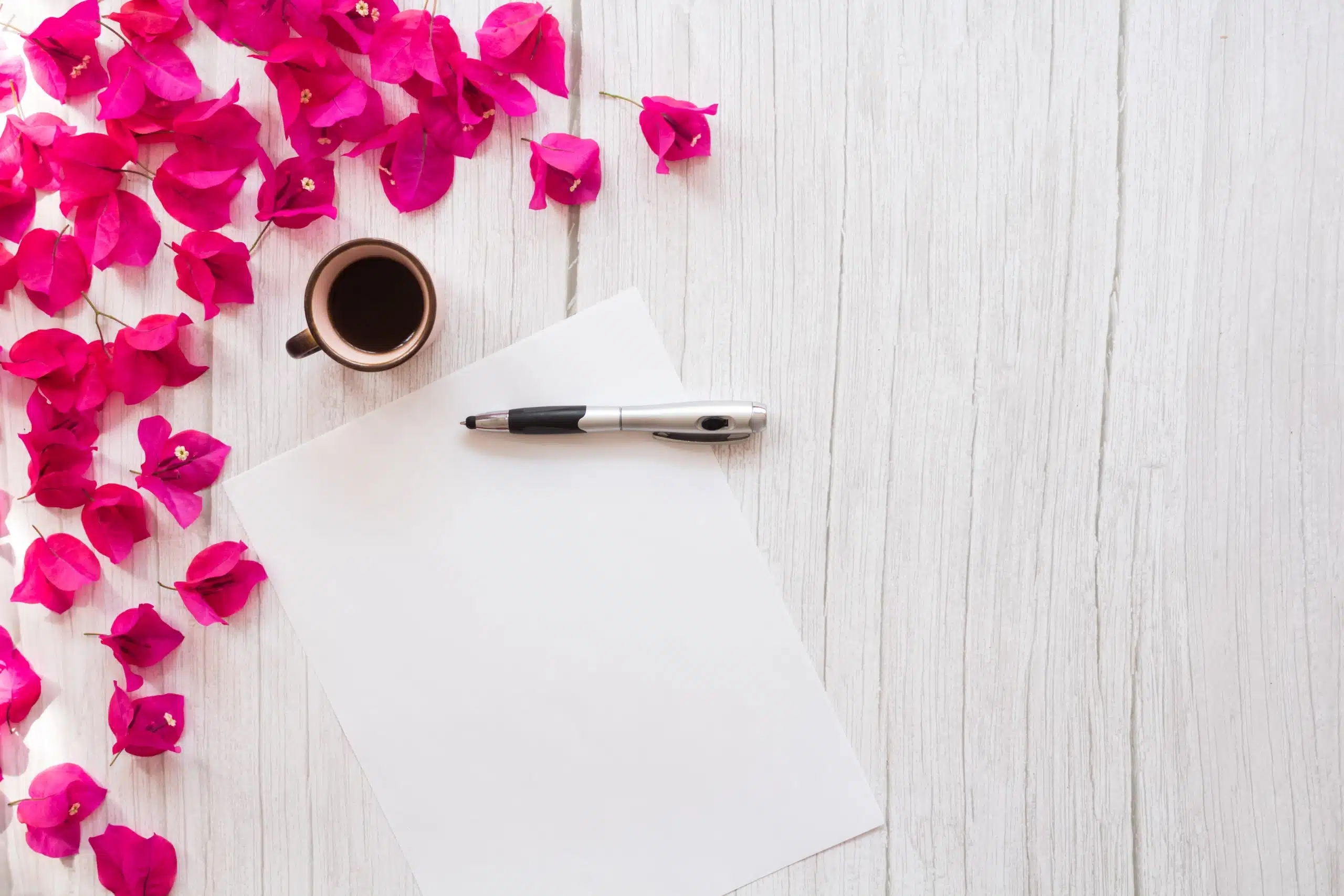 White paper with pen and a cup of tea, with bougainvillea flower petals on white wooden platform.