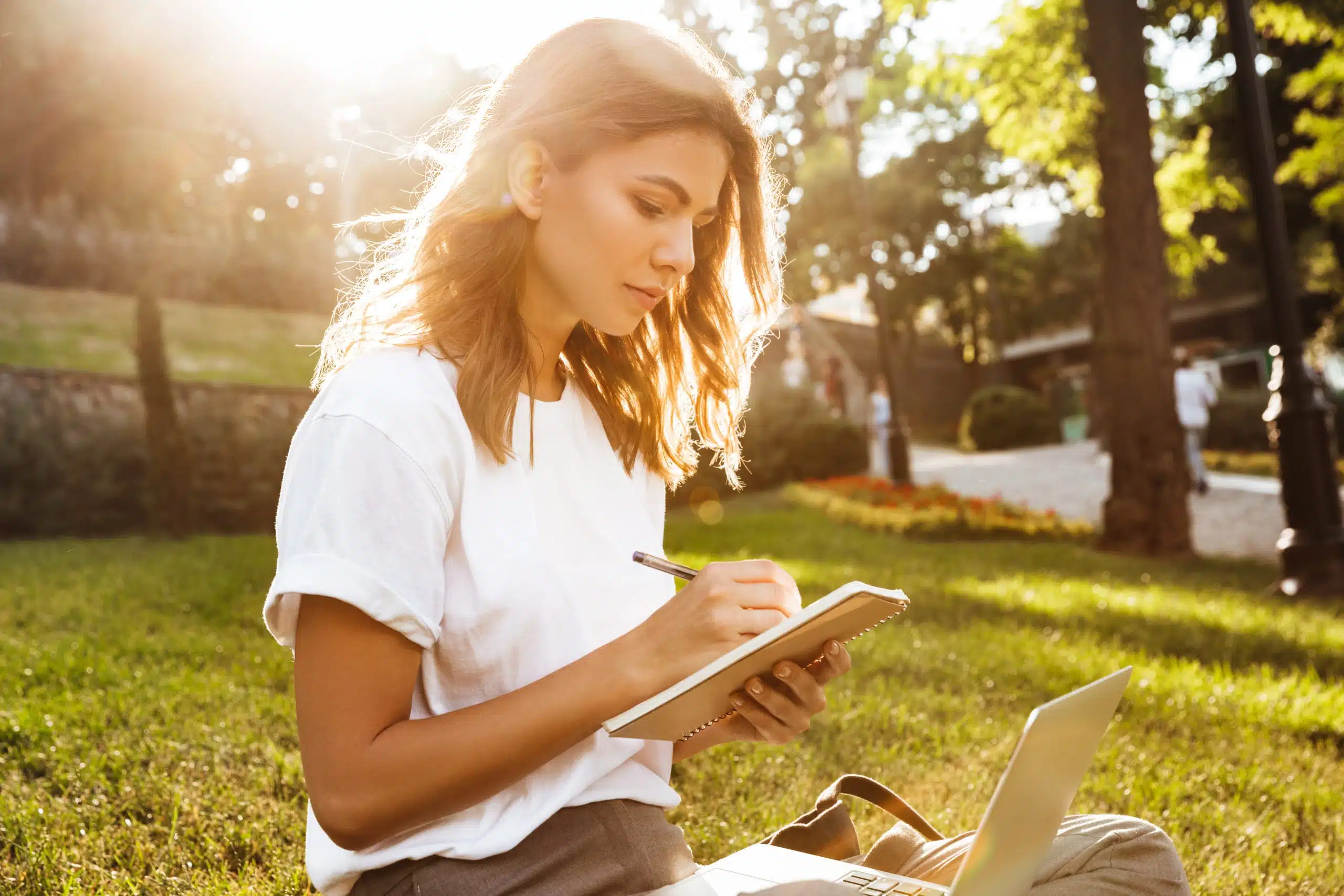 Young woman sitting on green grass in the park sunlit writing in her notebook.