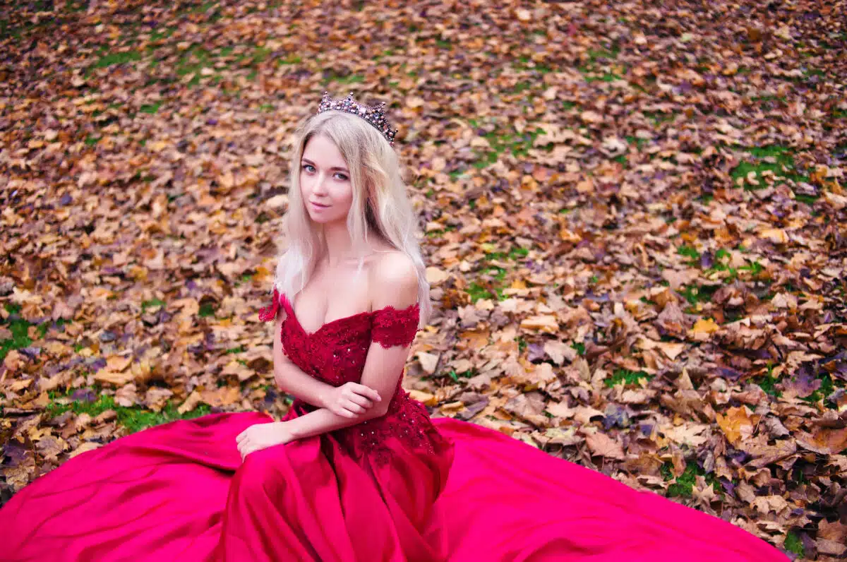 Princess with crown sitting in red dress on autumn leaves background. Queen