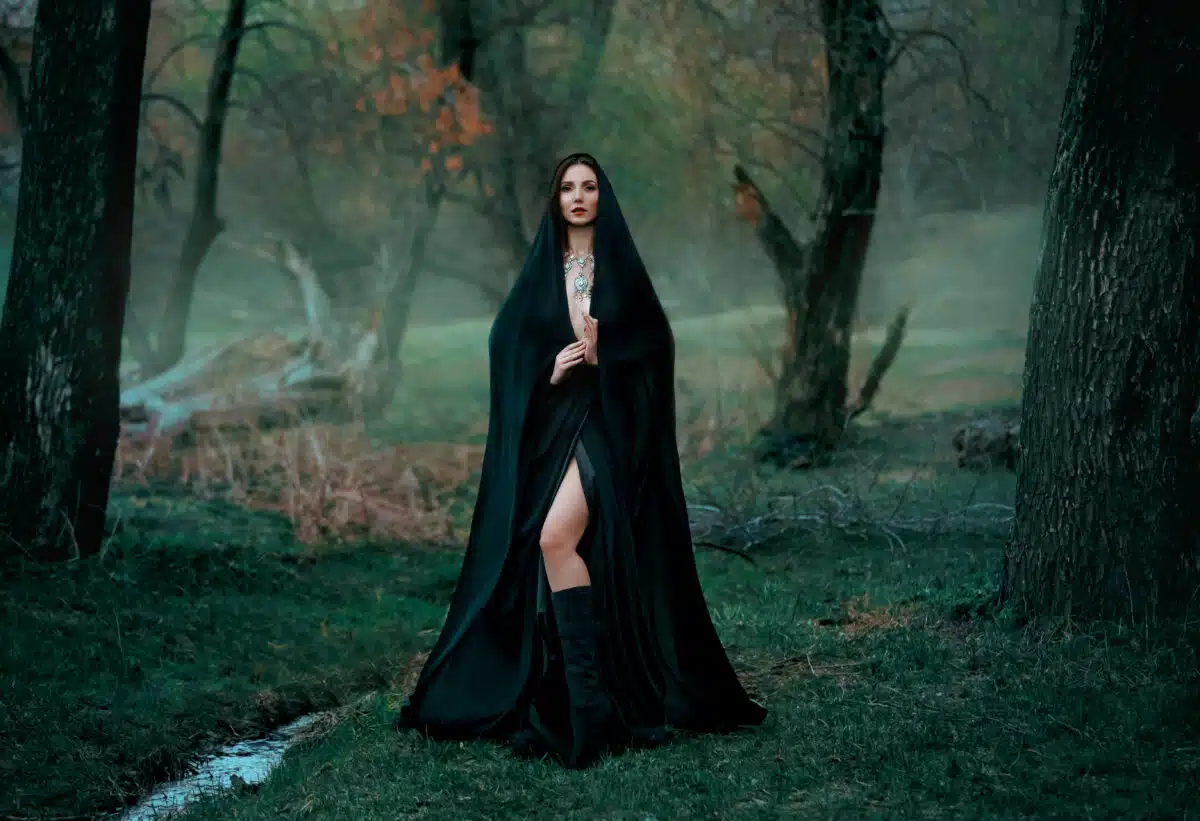 Mysterious fantasy gothic woman dark witch obsessed by evil. Girl demon vampire in black dress cape hood. walk in dark dense deep forest background, trees. Medieval queen in silk cloak, scarf posing.