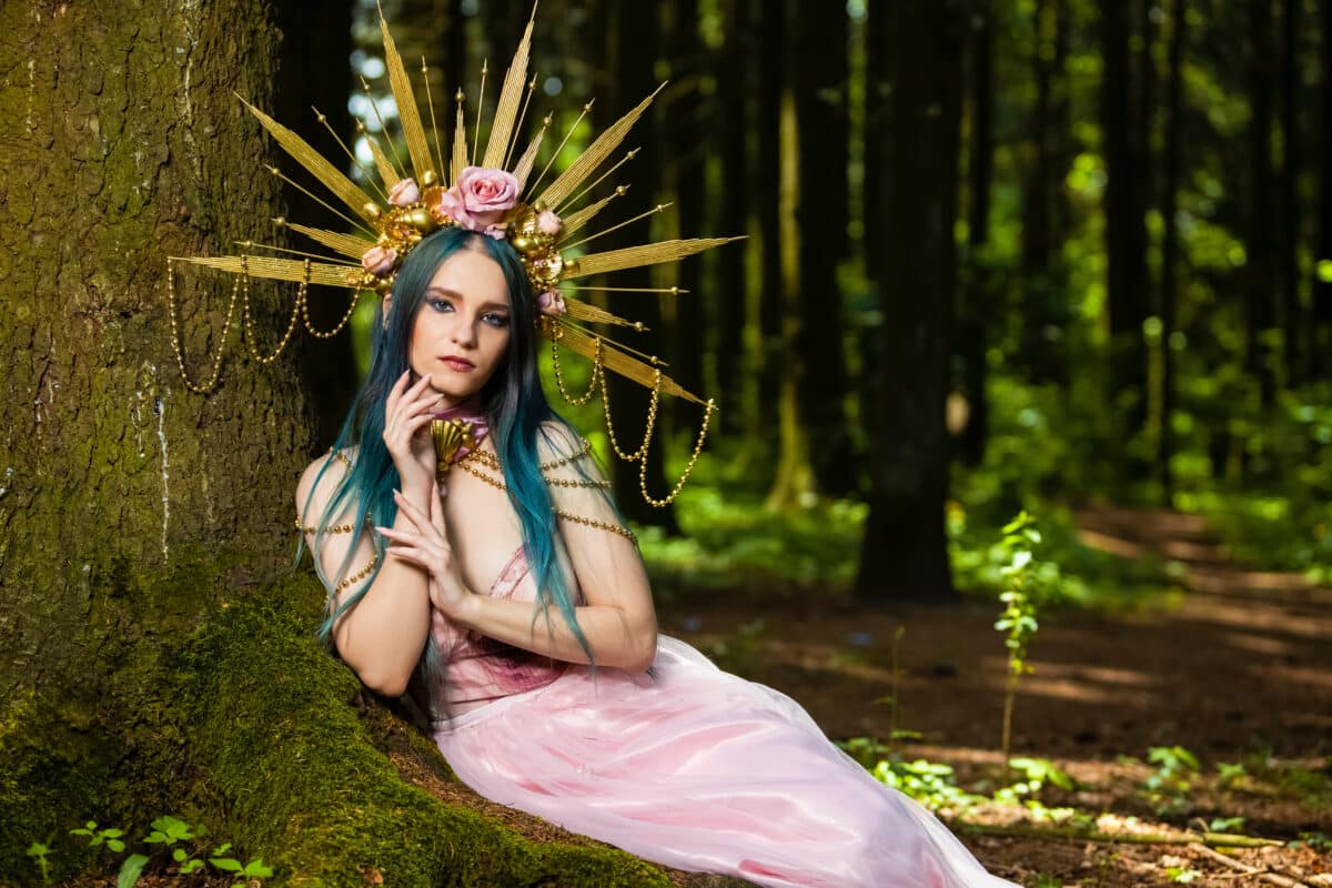Costume Play. Magnificent Crowned Forest Nymph with Flowery Golden Crown Posing in Summer Empty Forest Near Tree Stem.