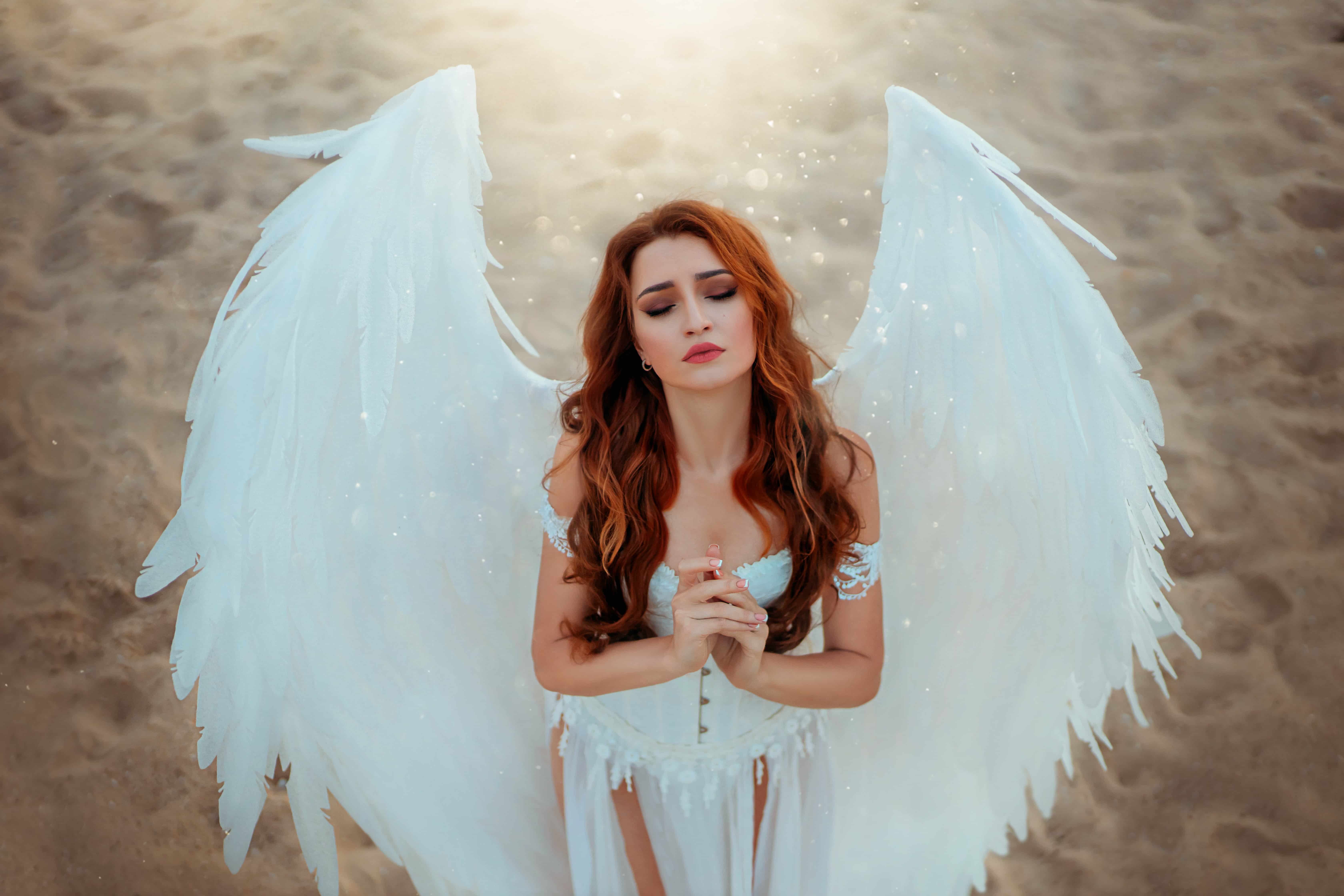 Fantasy woman in angel costume with white fake bird wings with artificial feather. Girl goddess with hands folded in prayer, closed eyes. Nature background beach, sand desert Divine blessing sun light.