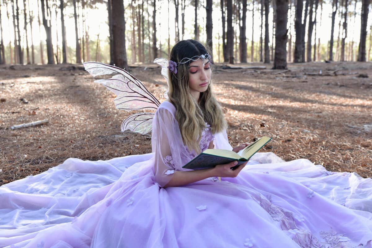 Full length portrait of beautiful young blonde model wearing a purple princess fantasy ball gown with flower crown diadem. Sitting pose in pine tree forest location background with golden afternoon