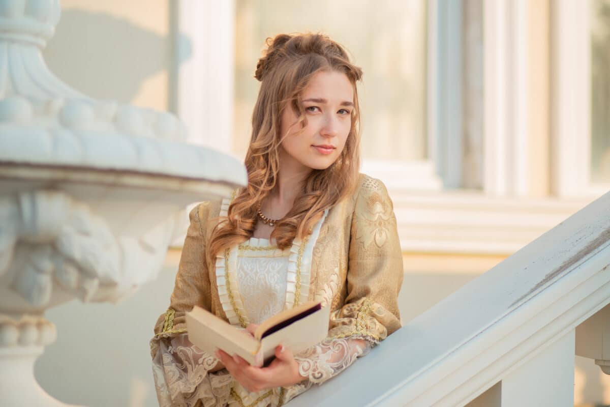 A beautiful young woman in an eighteenth century historical gold dress stands on the stairs of a mansion with a book in her hands. the princess is reading in the palace.