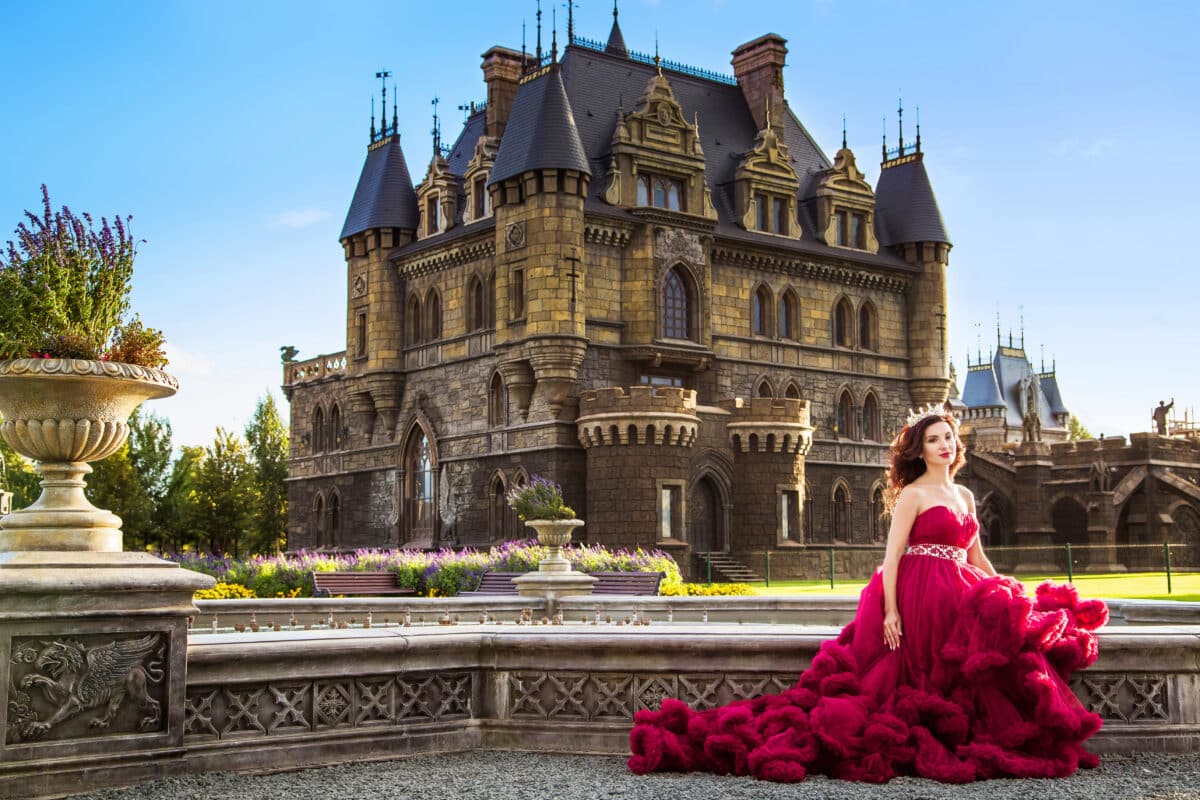 A beautiful woman, a princess in a red dress, sits by the fountain in a blooming garden. An ancient castle in the background. Medieval fantasy, European palace