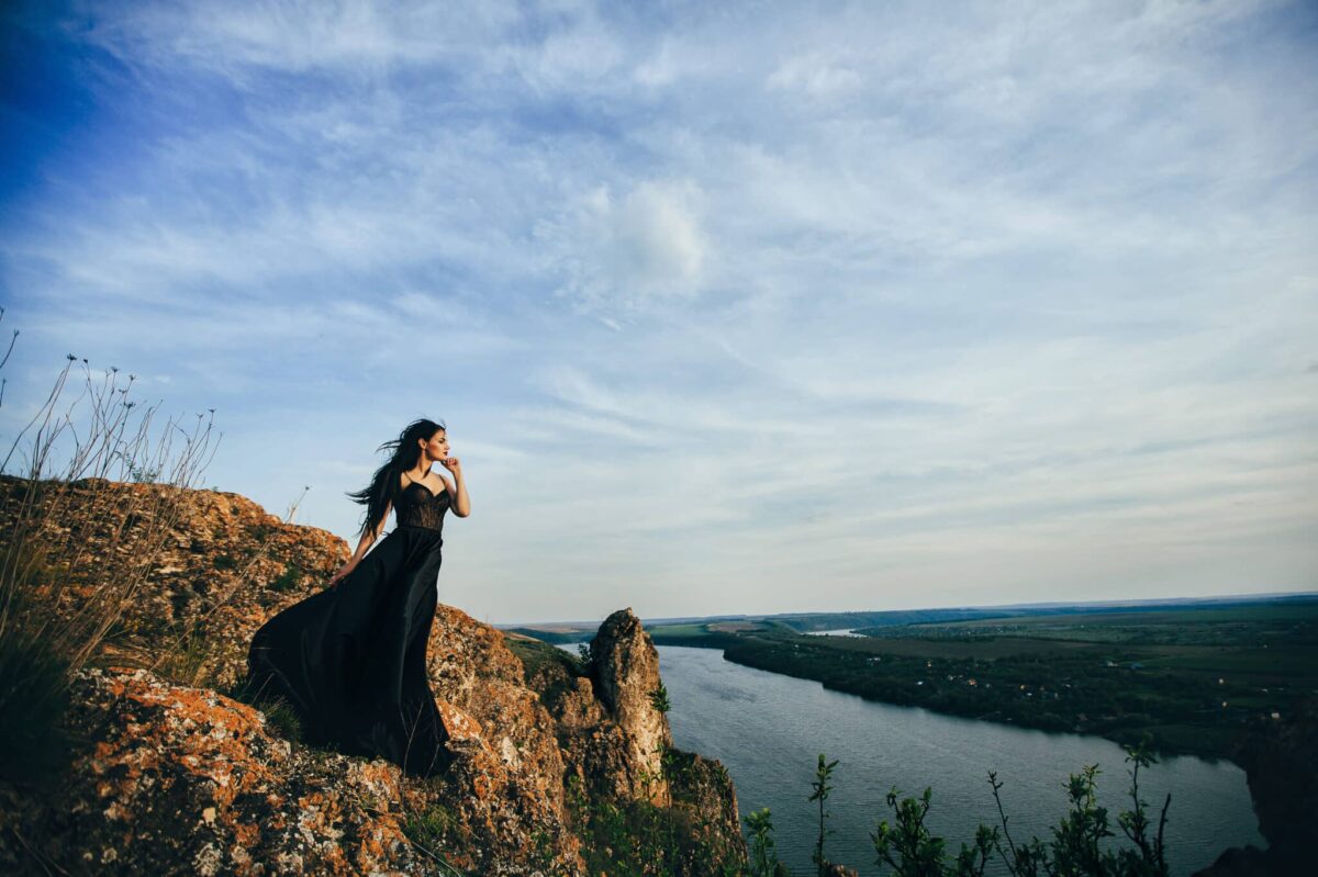 a lady dressed in a long black dress standing on a rock on a mountain by the river