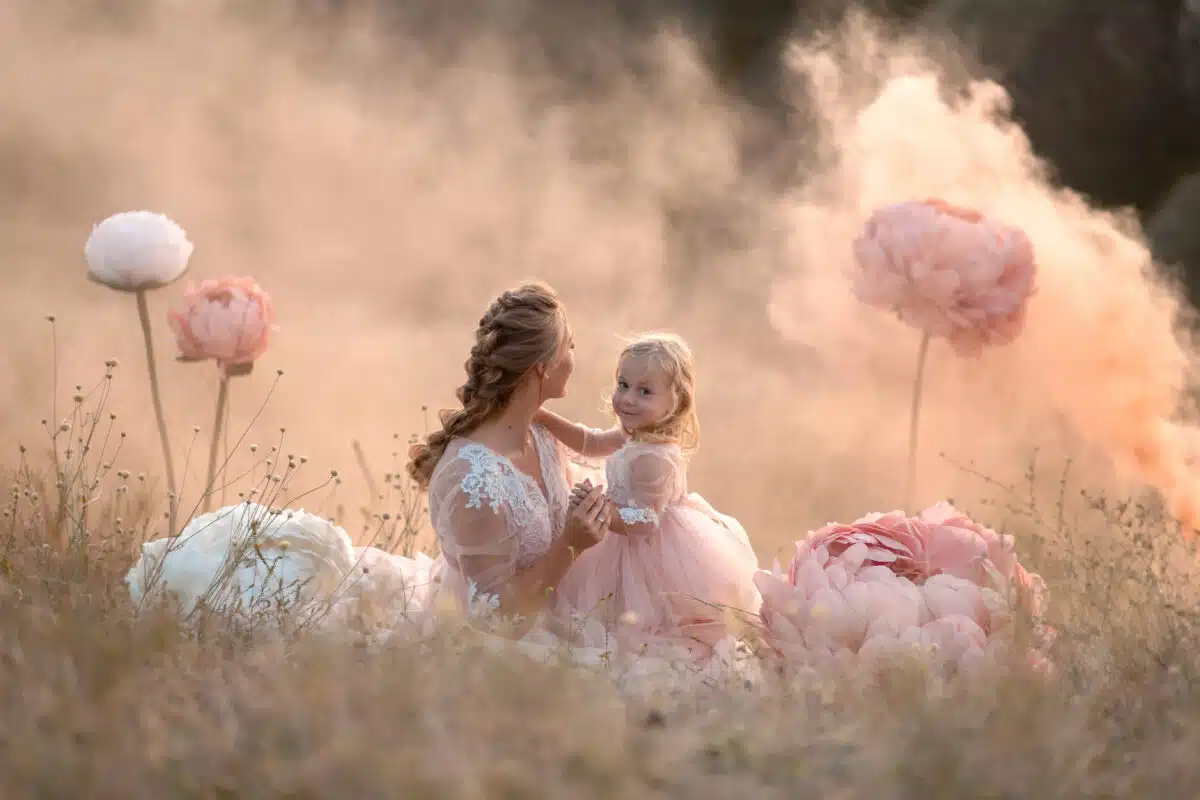 A little girl hugs mom sitting in a field surrounded giant pink peonies
