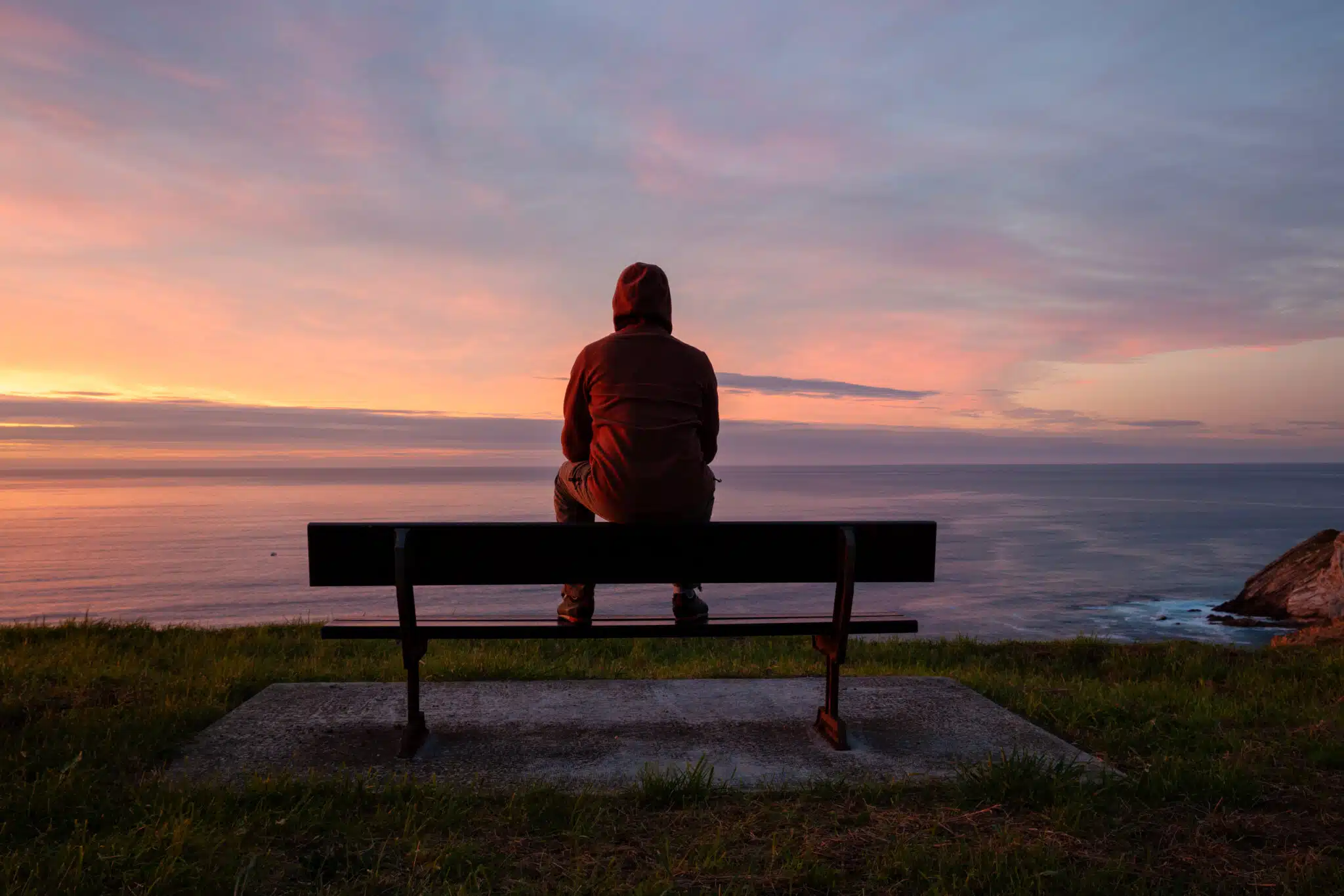 Lonely man sits alone on the rocky coast and enjoying sunset. View over rocky cliff to ocean horizon