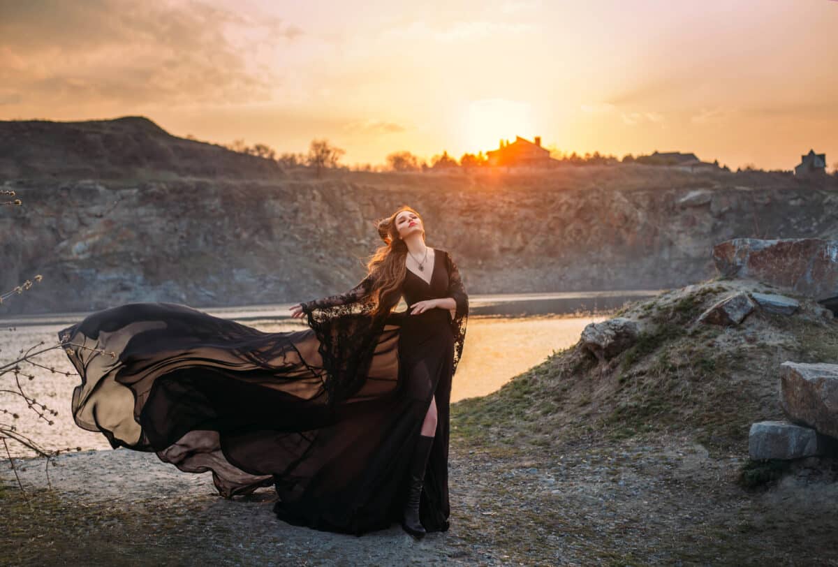 Fantasy sorceress in black fluttering silk dress with horns on her head. Dark gothic fashion glamorous character woman queen enjoying fabulous scenery of autumn nature. Sunset with warm rays sunshine