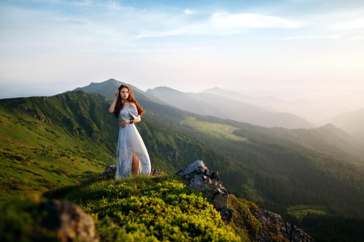 Young woman in long white dress. Beautiful young long hair woman posing in high mountain scenery. Woman standing on mountain top and contemplating the sunset