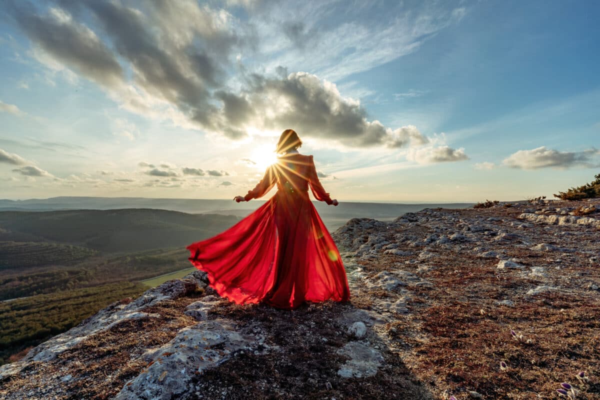 Woman in red dress stands on the mountain with a magical view of the sky, clouds and sunrise