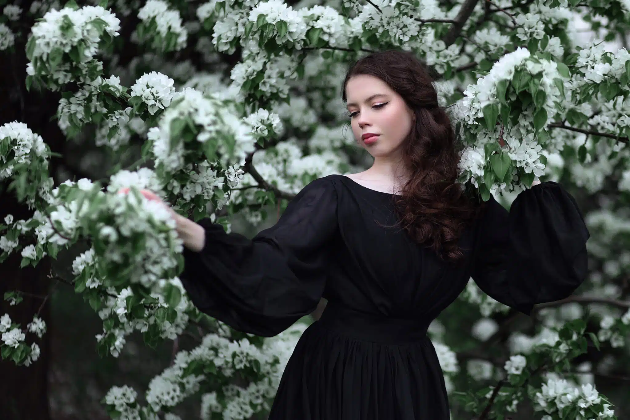 melancholic young woman in black dress standing among blooming trees
