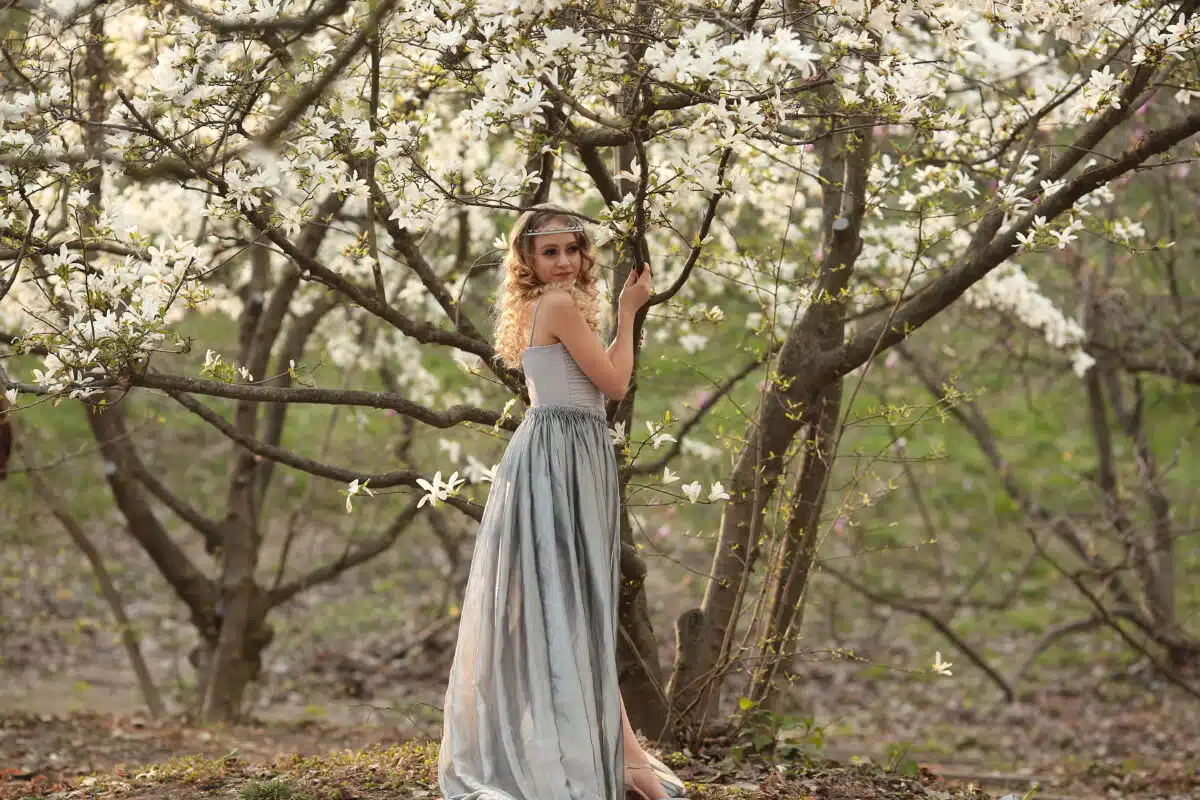 young beautiful blonde in a silver dress stands among blooming trees