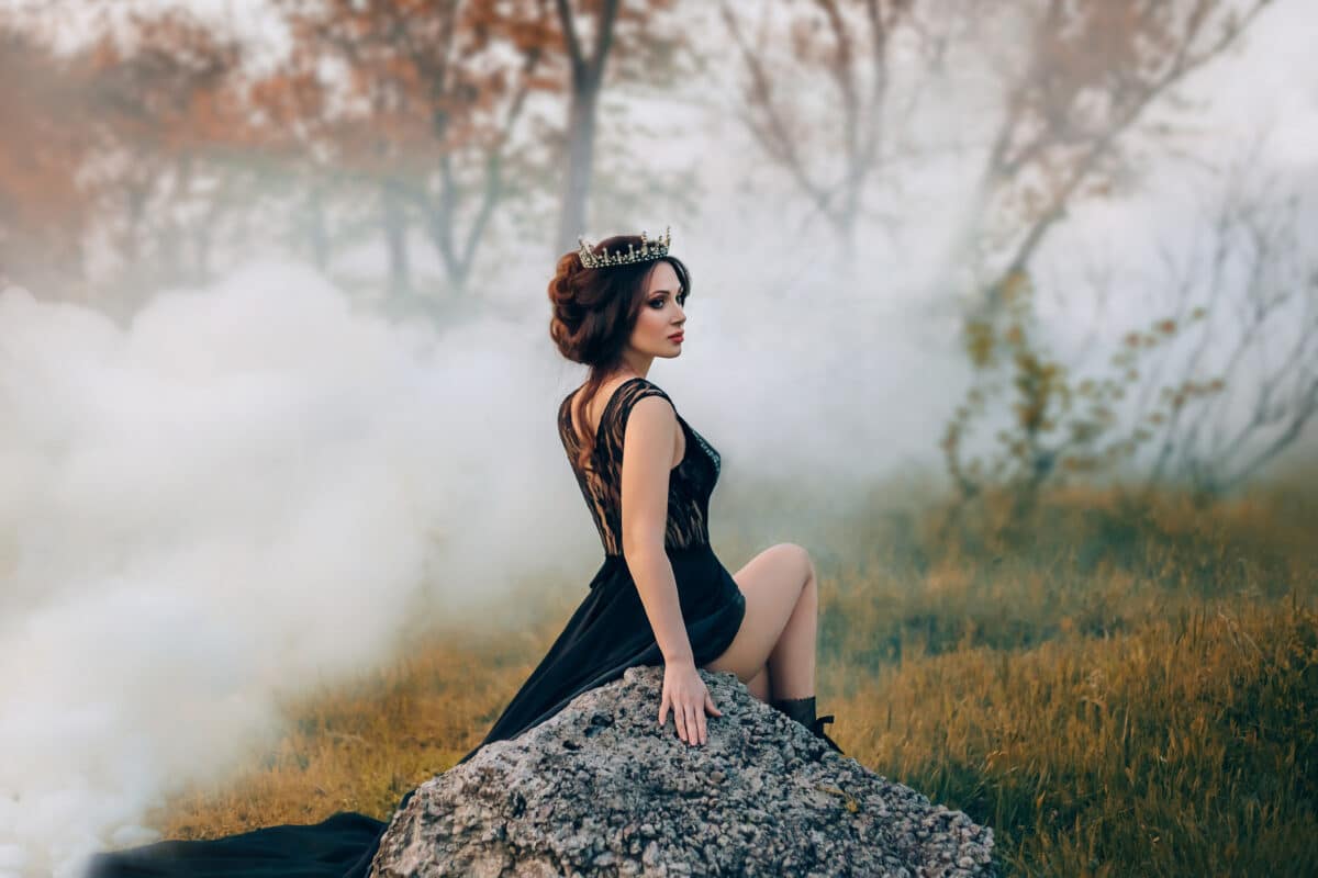 The majestic lady, the dark queen, sits on the stone baring her leg. The brunette girl in the gothic crown. The background is golden autumn and thick fog, clouds of smoke.