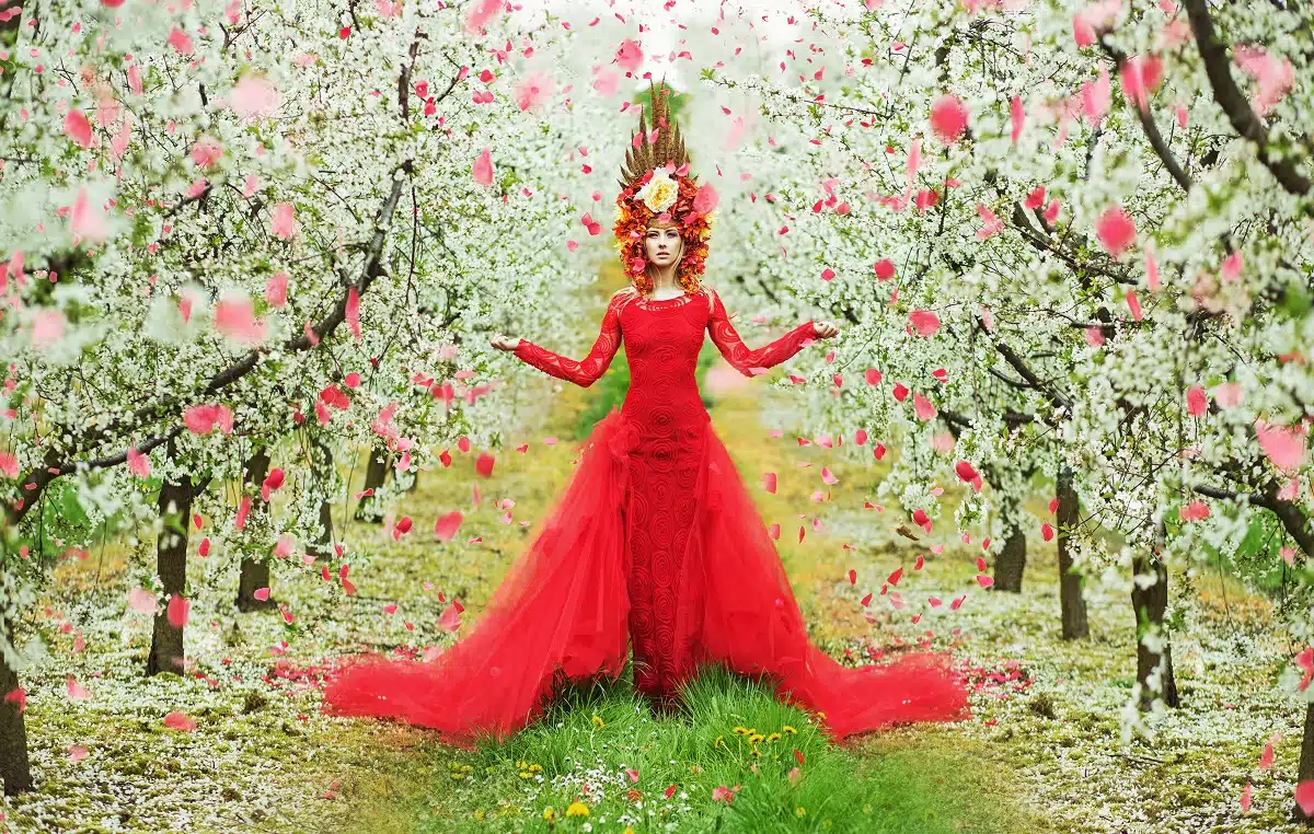 Lady of spring in a majestic red dress walking during the petal rain in the orchard