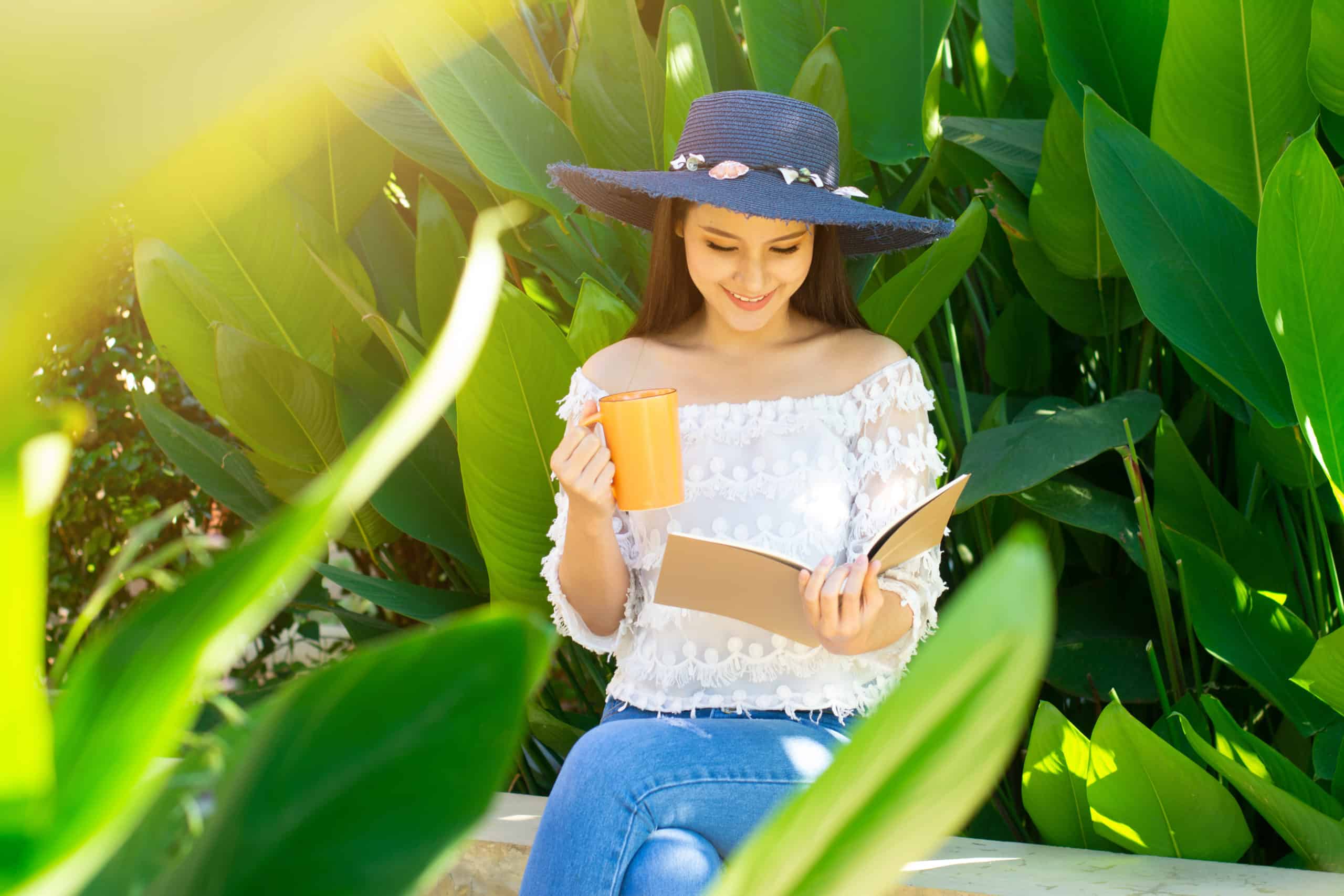 Young asian woman reading a book in the garden and holding cup.