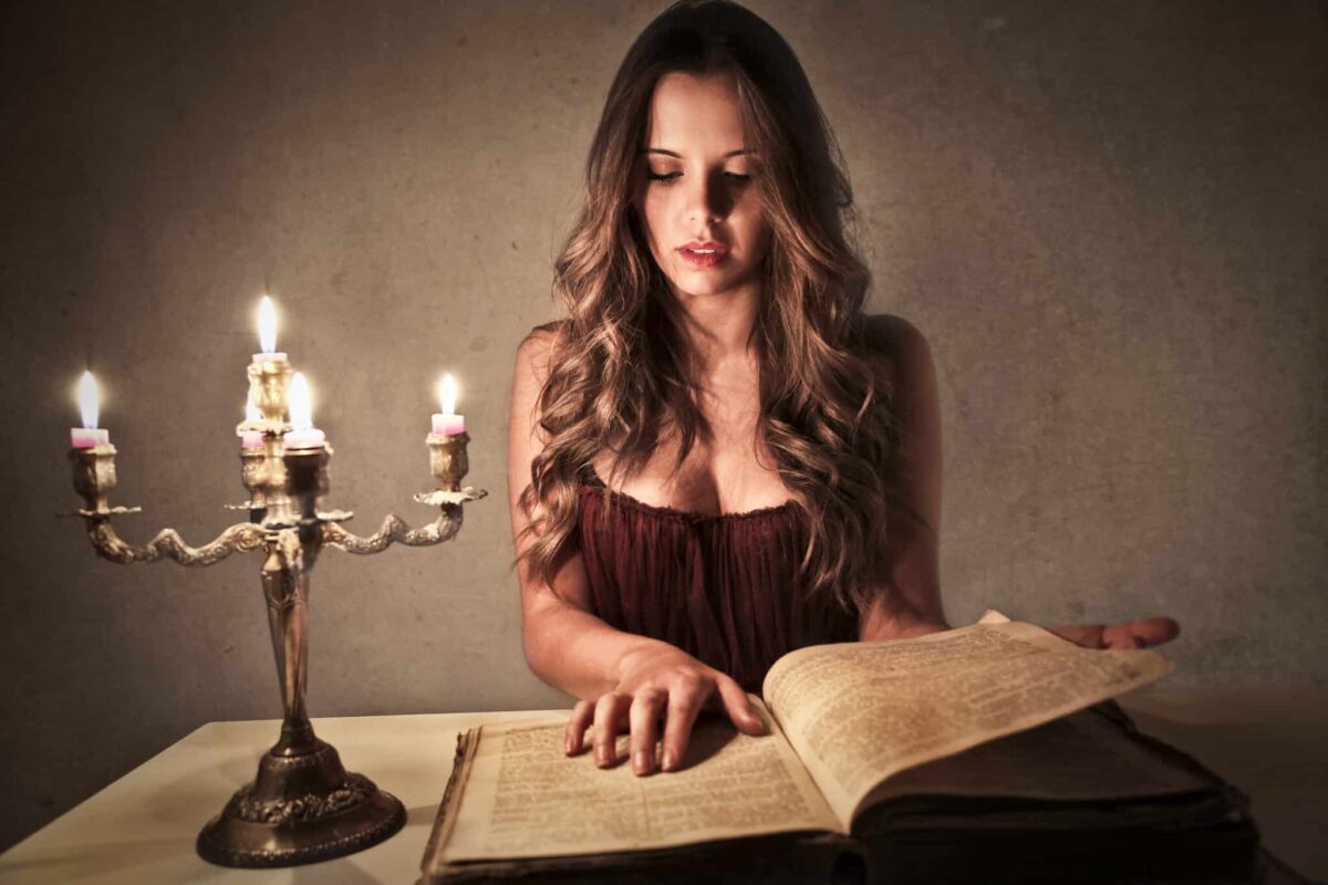 a woman reading a big vintage book at a wooden desk with lit candles