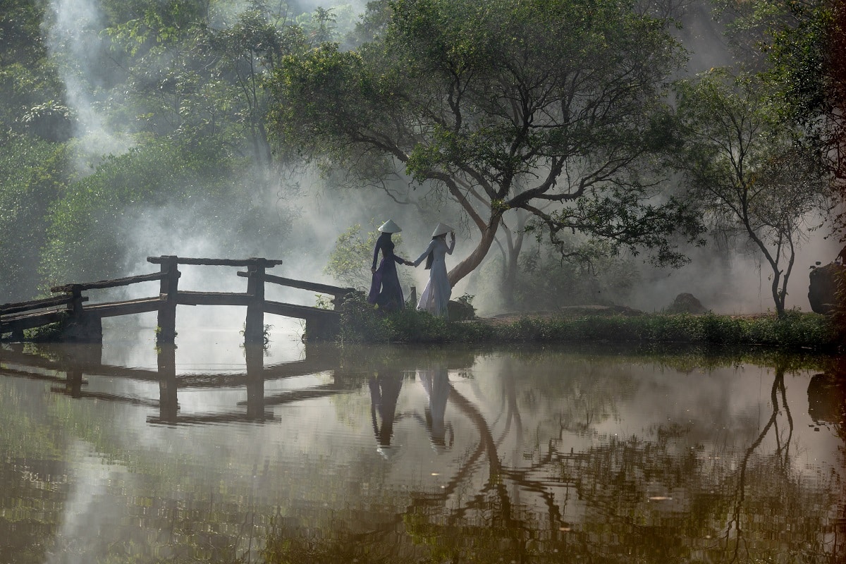 Vietnamese ladies wearing traditional dresses and hats cross the beautiful traditional bridge over the misty lake
