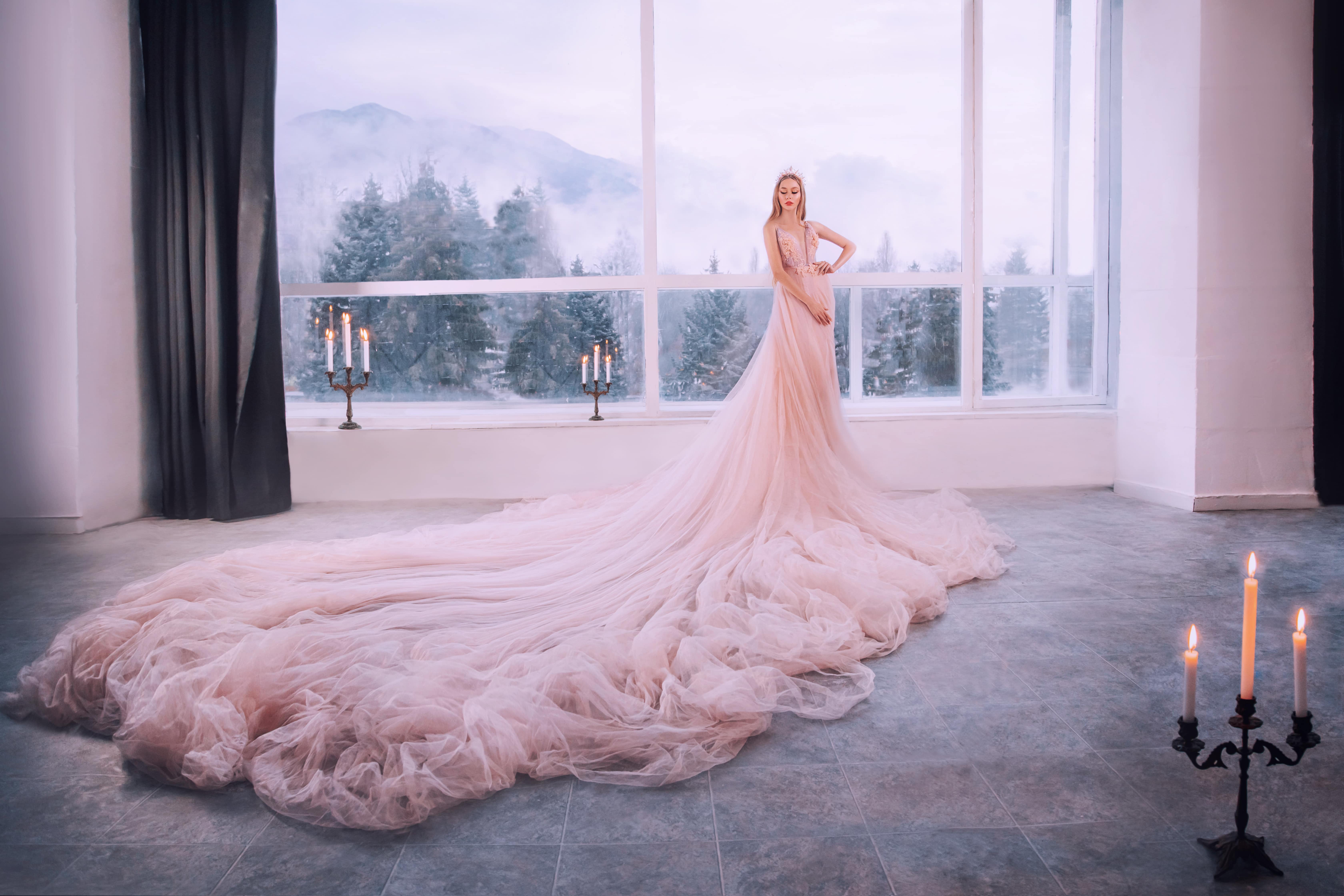 Fantasy girl princess in luxurious pink fluffy lush dress stands in castle room by vintage window with winter nature, forest trees mountains. Woman queen bride in wedding dress, long train, hem skirt