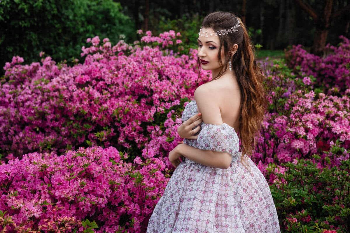 spring outdoor portrait of a beautiful sexy brunette woman with curly hair, in a long light dress and in a crown, stands in flowers among the branches of pink azalea bloom