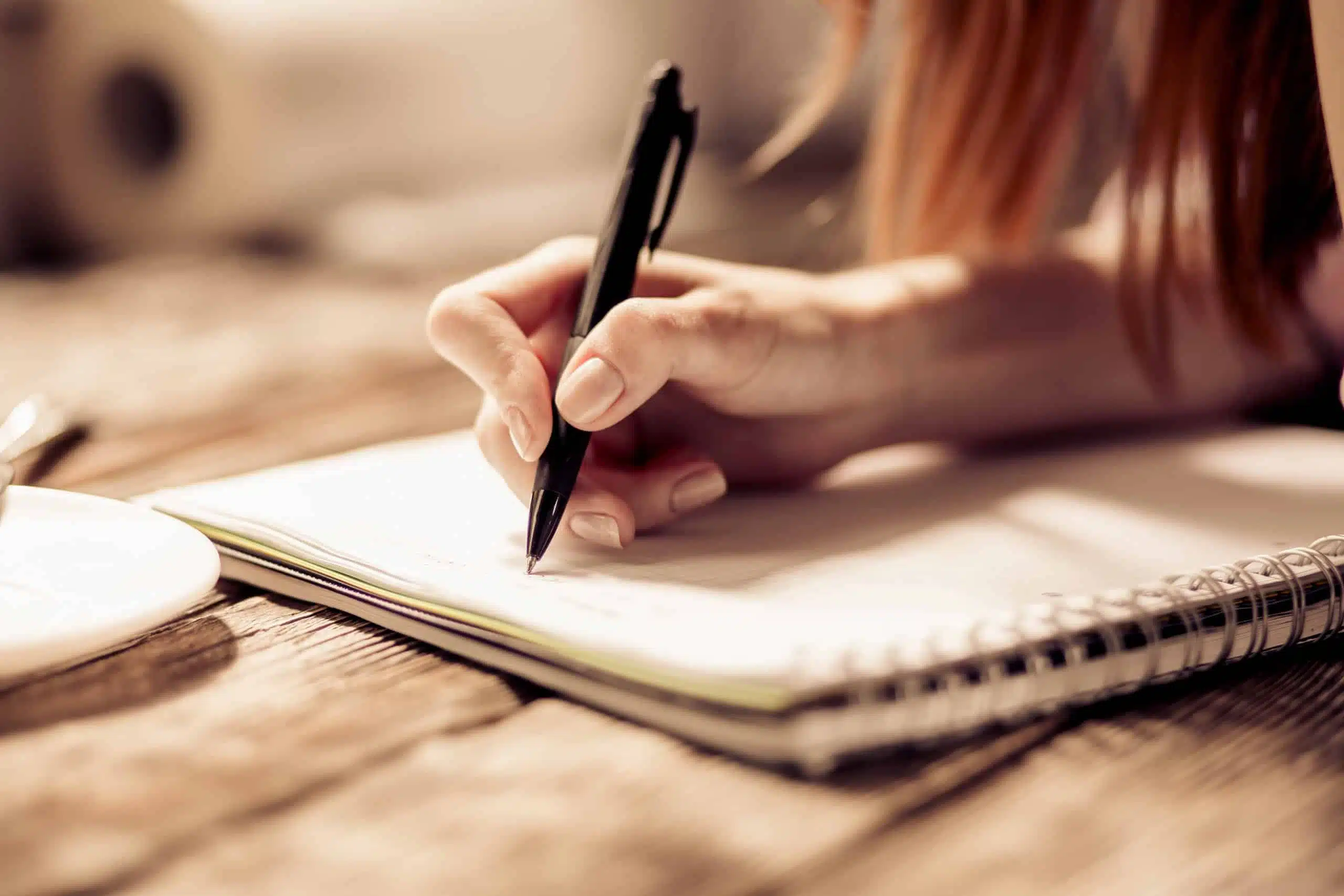 Woman writing with pen on notebook on wooden table.