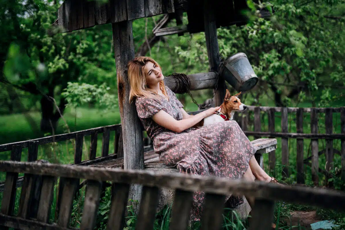 a blonde village girl in a dress and brown shoes is sitting on a wood pit with her dog, a green garden behind her