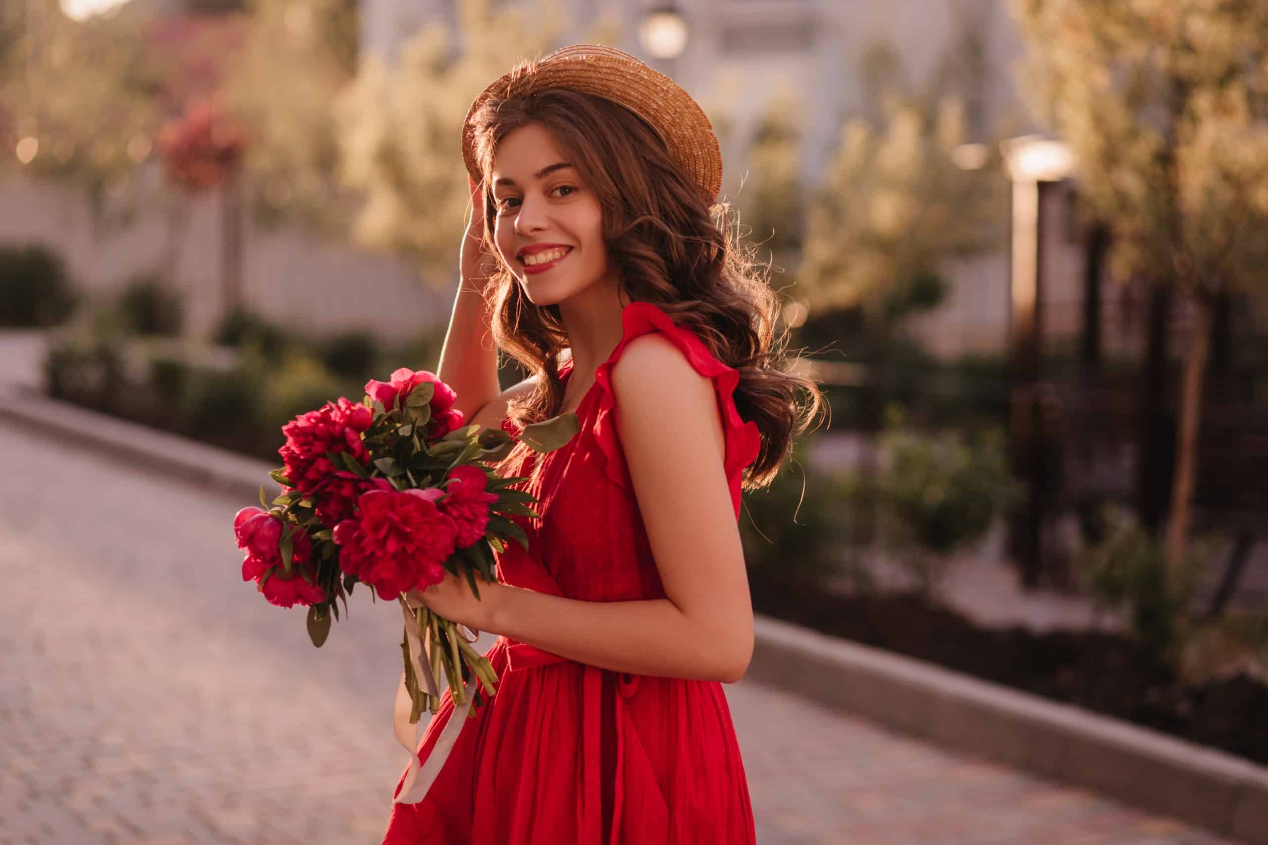 A lovely girl in red is walking on the street with a boquet of red roses in a romantic mood before the sunset