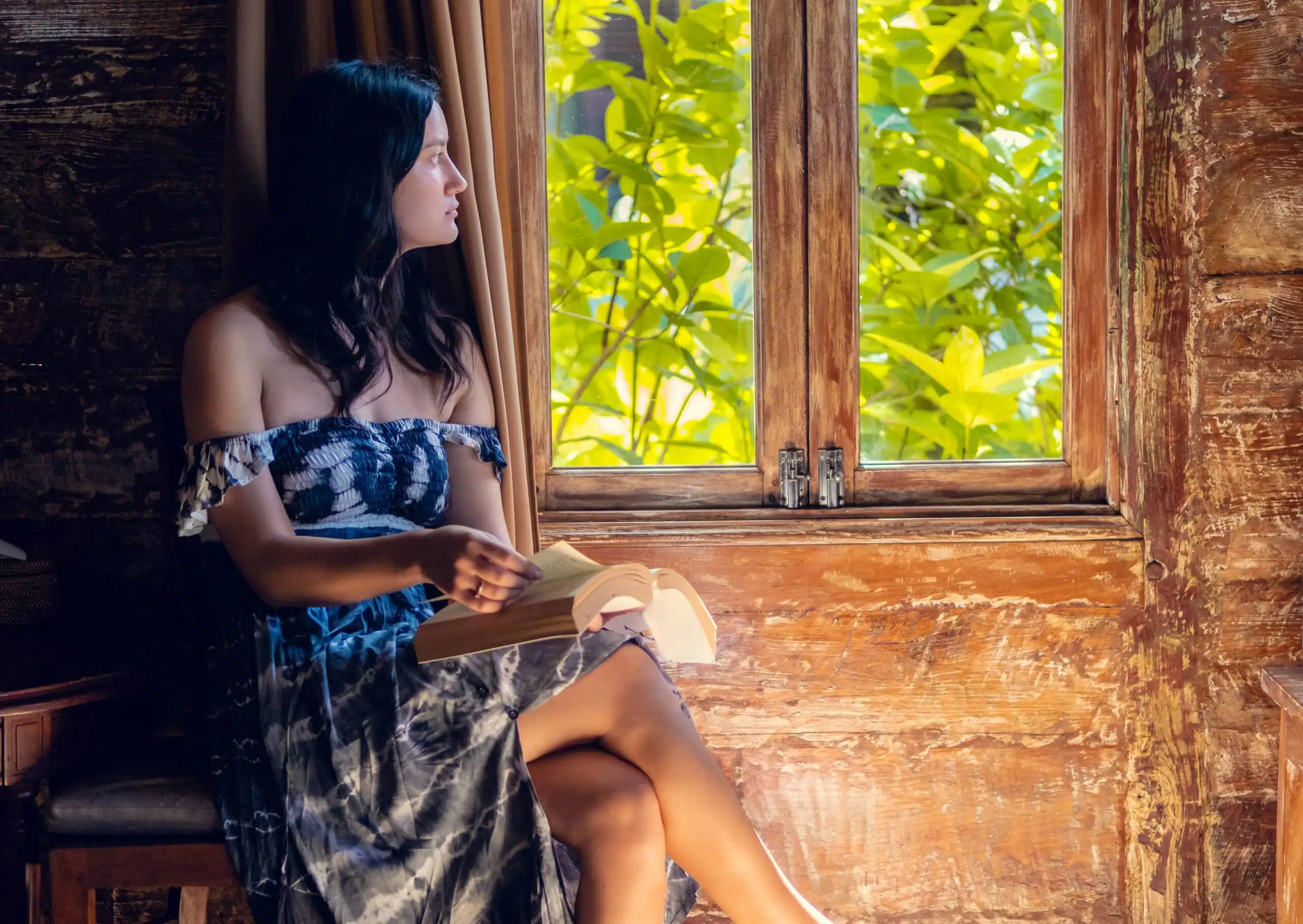 Beautiful pensive young woman sitting by the window in wooden bunglow rustic house and reading a book
