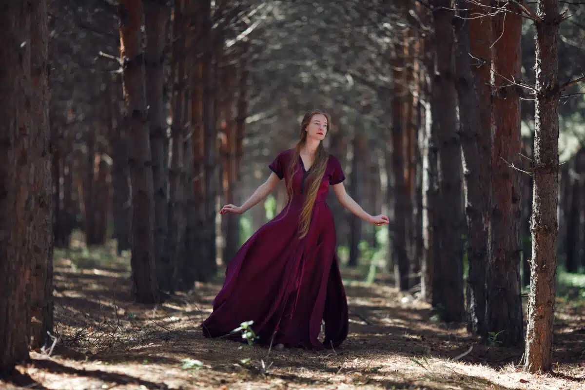 A tall, slender woman with long blond hair walks in the woods in a burgundy dress
