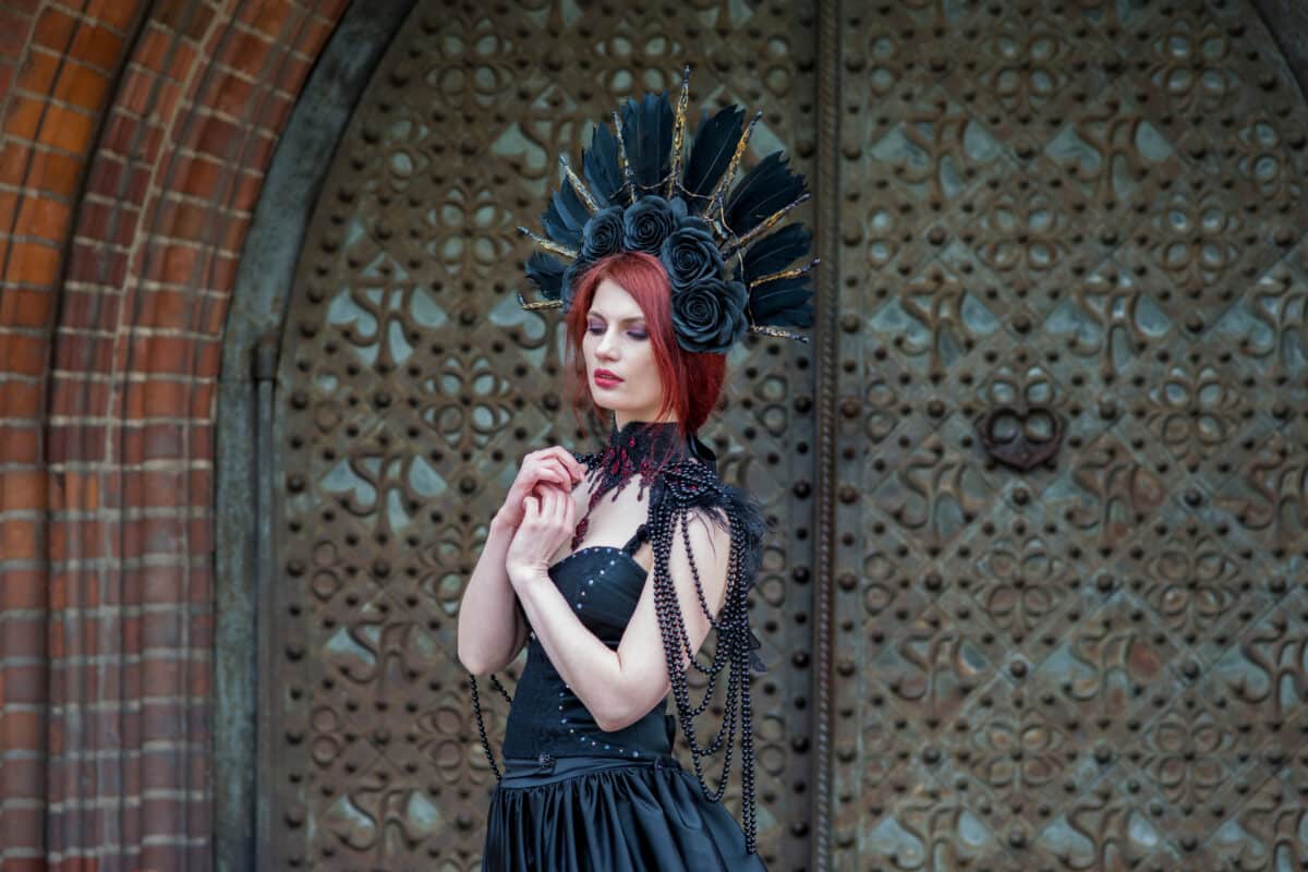 Fashionable Gothic Woman in Long Black Dress. Wearing Artistic Feather Crown. Posing Against Old Castle Gates.