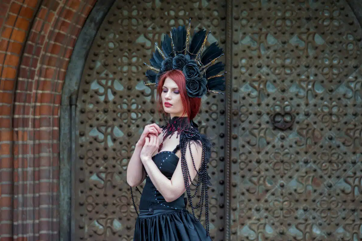 Fashionable Gothic Woman in Long Black Dress. Wearing Artistic Feather Crown. Posing Against Old Castle Gates.
