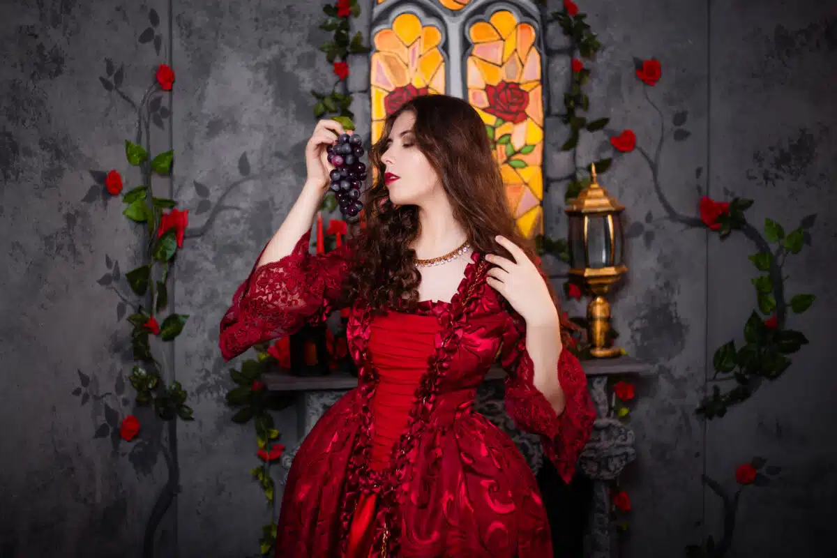 A beautiful girl in a magnificent red dress of the Rococo era stands against a fireplace, a window and flowers with a bunch of grapes in her hands.