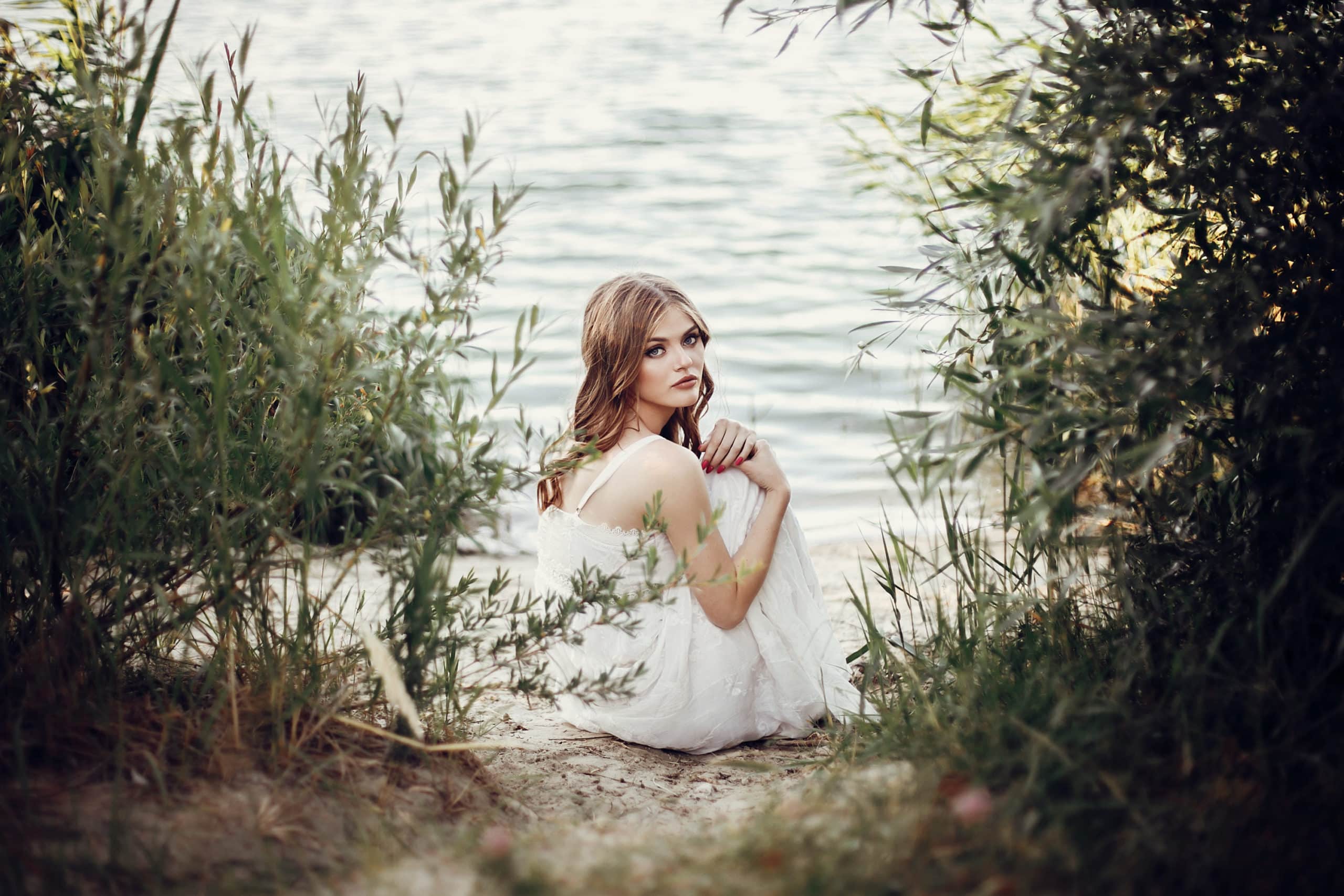 Beautiful boho girl sitting on sandy lake behind the bushes.